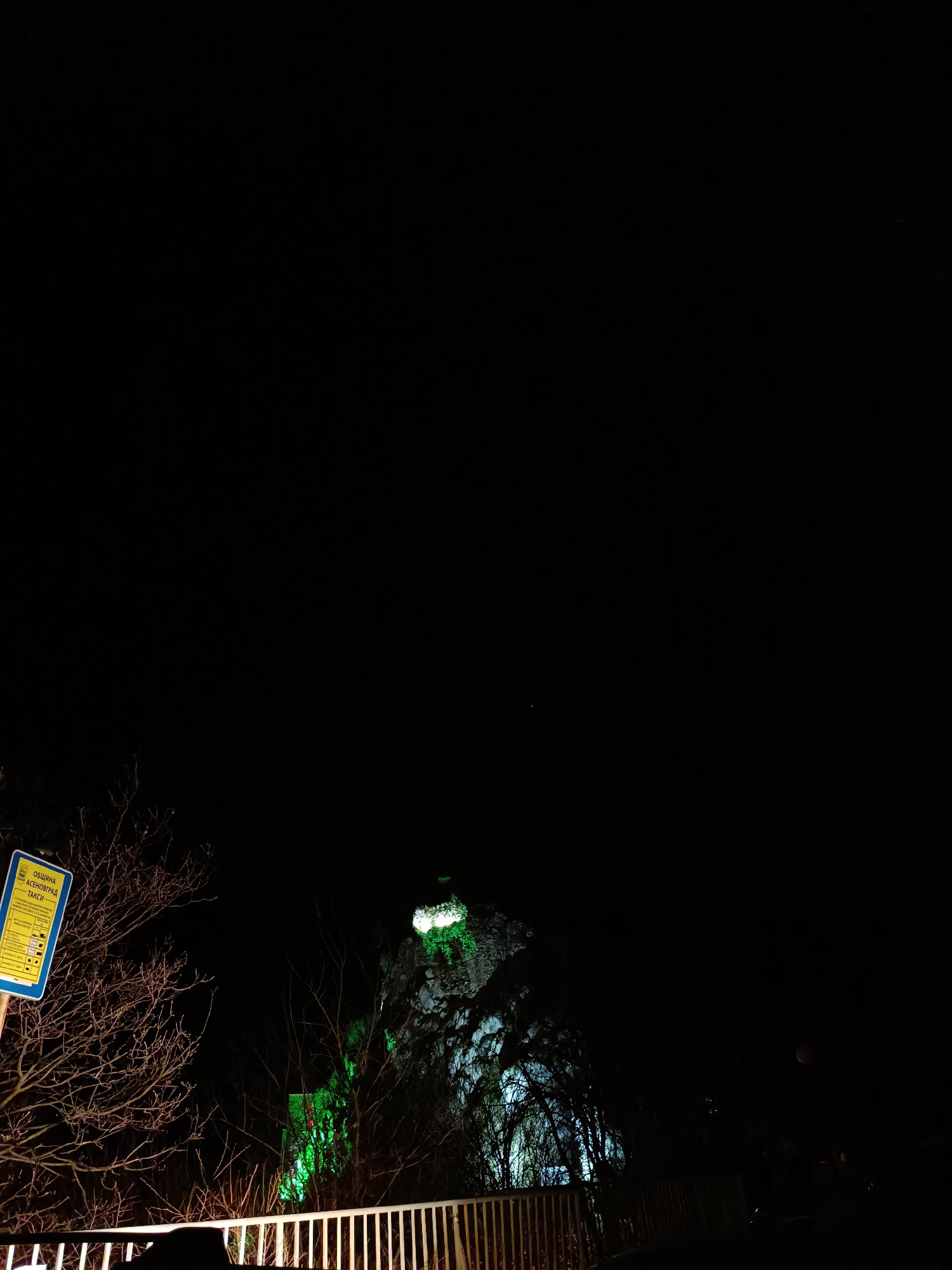 The image displays a nighttime scene in Asenovgrad, Bulgaria. A prominent structure, identified as Asen's Fortress, is visible atop a dark, rocky hill in the upper-center, illuminated by an artificial green light source. Below the fortress, a cluster of deciduous trees with bare branches partially covers the lower rock face and hillside. Some of these trees are also illuminated with green light, while others further to the right show faint blue-white illumination.
In the lower-left foreground, a rectangular blue municipal sign is partially visible. The sign features yellow text in Cyrillic, clearly reading "ОБЩИНА АСЕНОВГРАД ТАКСИ" (Municipality of Asenovgrad Taxi), along with several small pictograms. A white metal railing or barrier extends horizontally across the lower foreground of the image, illuminated from the left by an unseen light source. The sky above is completely dark and featureless. No individuals or vehicles are present in the scene.