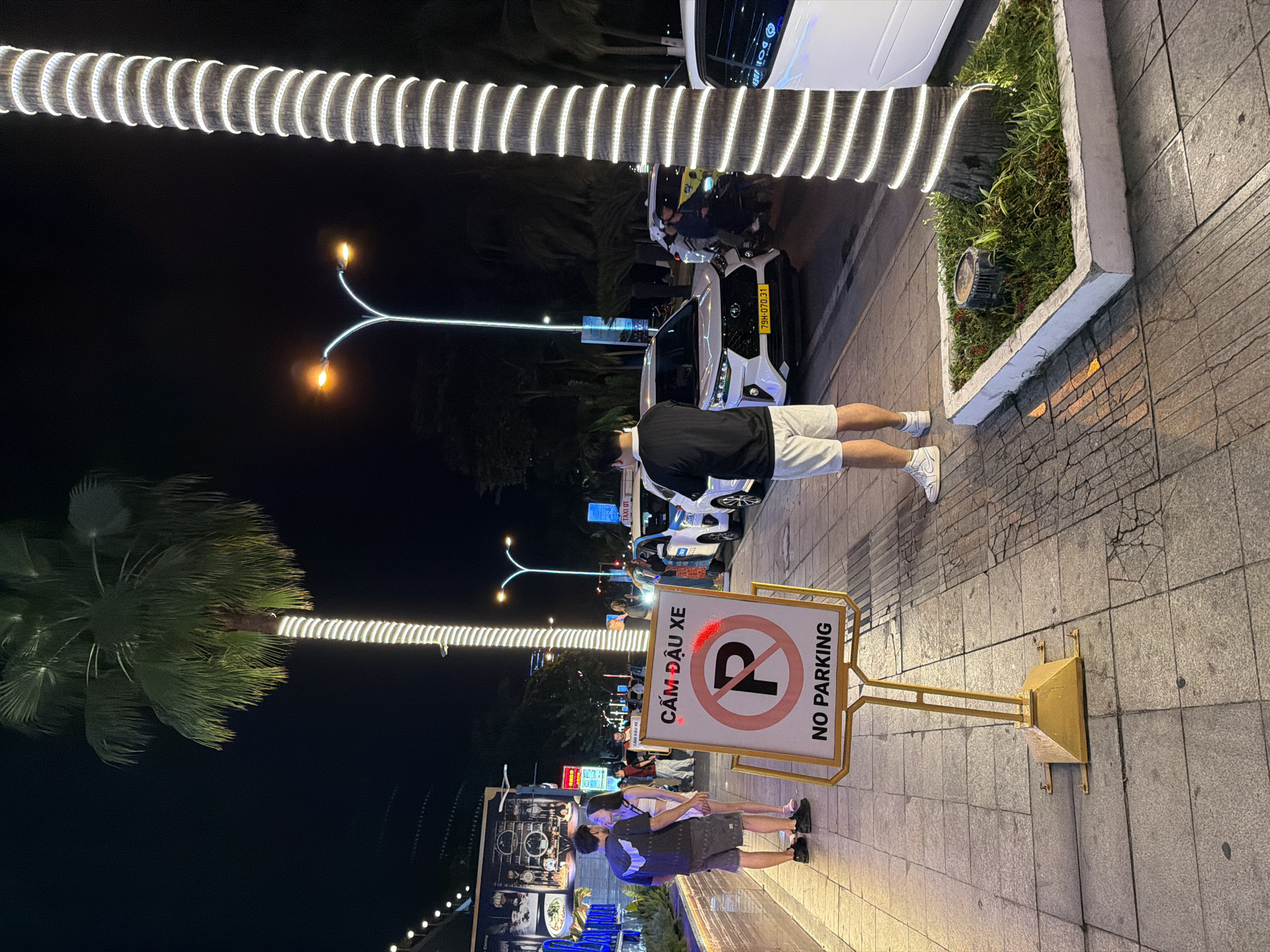 In the city of Nha Trang, Vietnam, at night, a street scene unfolds with a prominent "No Parking" sign in the foreground. The sign, a circular red symbol with a diagonal line through a "P," is in Vietnamese and English. Behind the sign, a man stands facing away from the camera, looking at the street. To his left, a couple stands close together, appearing to be in an embrace or about to kiss. They are on a sidewalk lined with palm trees adorned with white lights. Various vehicles, including a white taxi and other cars, are visible on the street. Buildings with illuminated signs are in the background, suggesting a commercial or entertainment district. The overall atmosphere is lively, typical of a nighttime street in a Vietnamese coastal city.