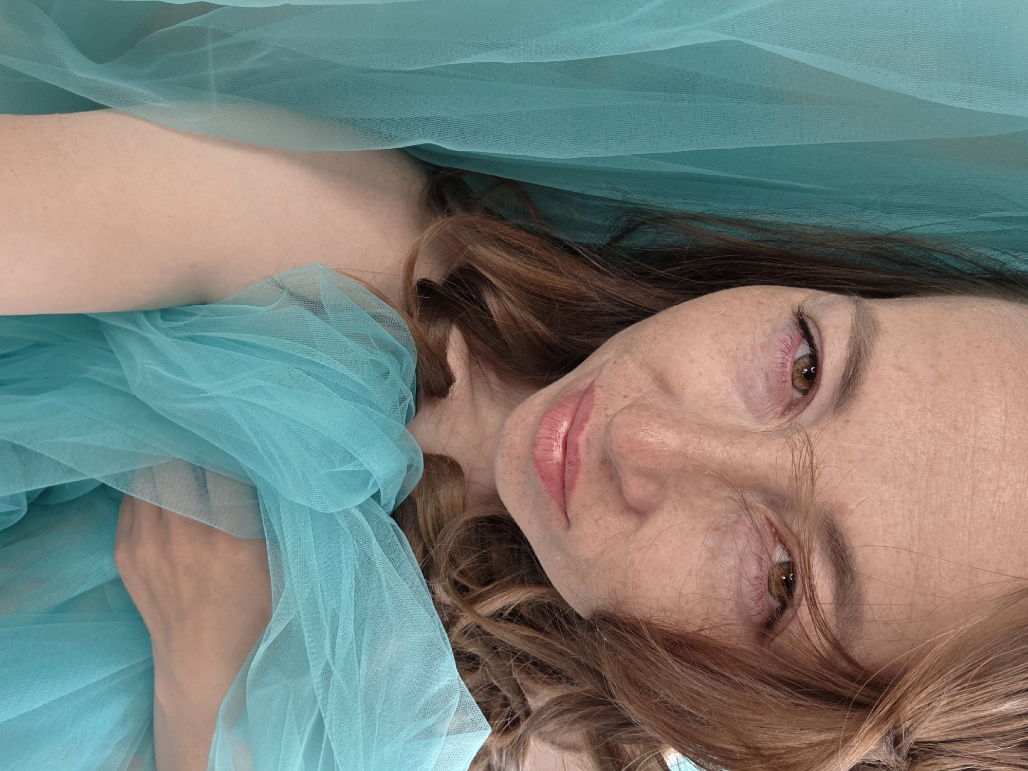 A woman with freckles and auburn hair is lying down, partially covered by a light blue sheer fabric. The image appears to be taken from a close-up, slightly overhead perspective. No information is available to determine if the location is Chaikovsky, Russia.