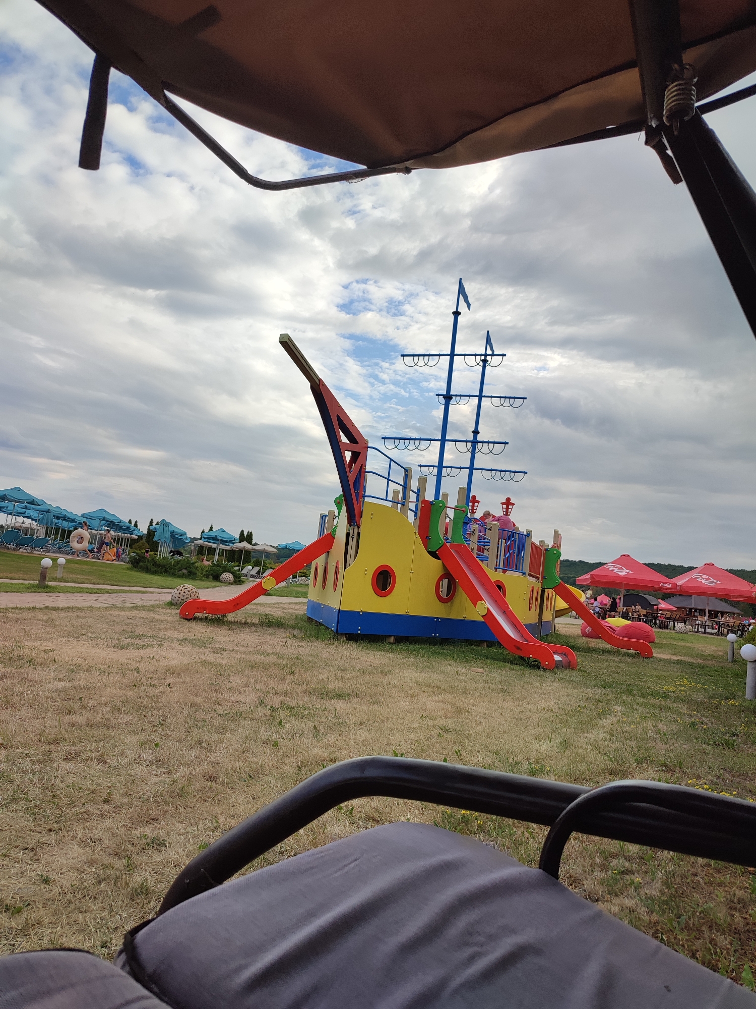 An outdoor shot in Shcherbani, Ukraine captures a colorful, ship-themed playground structure with red and yellow slides and blue masts, set on a dry, grassy field under a cloudy sky. In the background, rows of turquoise beach umbrellas and lounge chairs suggest a resort or recreation area. Several people are visible near the beach umbrellas, and a few are on the playground. The foreground shows the edge of a dark-colored seating area, possibly a swing or a shaded pavilion. Red Coca-Cola umbrellas are visible to the right, indicating a cafe or dining area.
