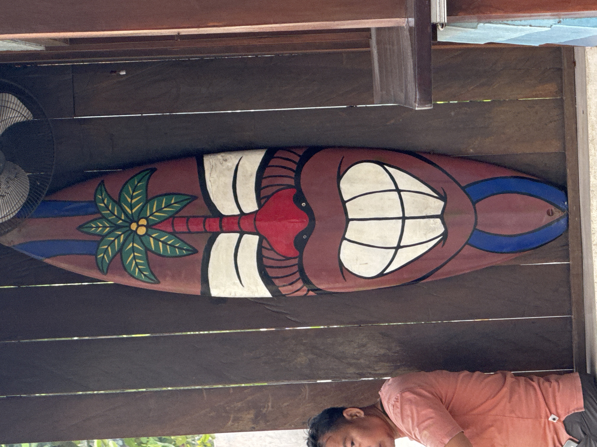 In Kuta, Indonesia, a vertical surfboard with a painted tiki face is displayed against a wooden slatted wall. The tiki face is predominantly brown with a red nose, large white teeth, and black outlines. A stylized palm tree with green fronds and yellow coconuts is painted at the top of the surfboard. To the left of the surfboard, a man wearing a coral pink t-shirt and dark pants is partially visible, facing to the right. The background consists of dark wooden slats, with some greenery visible through the gaps. A large industrial fan is mounted above the surfboard. A wooden counter or shelf is visible on the right side of the frame.