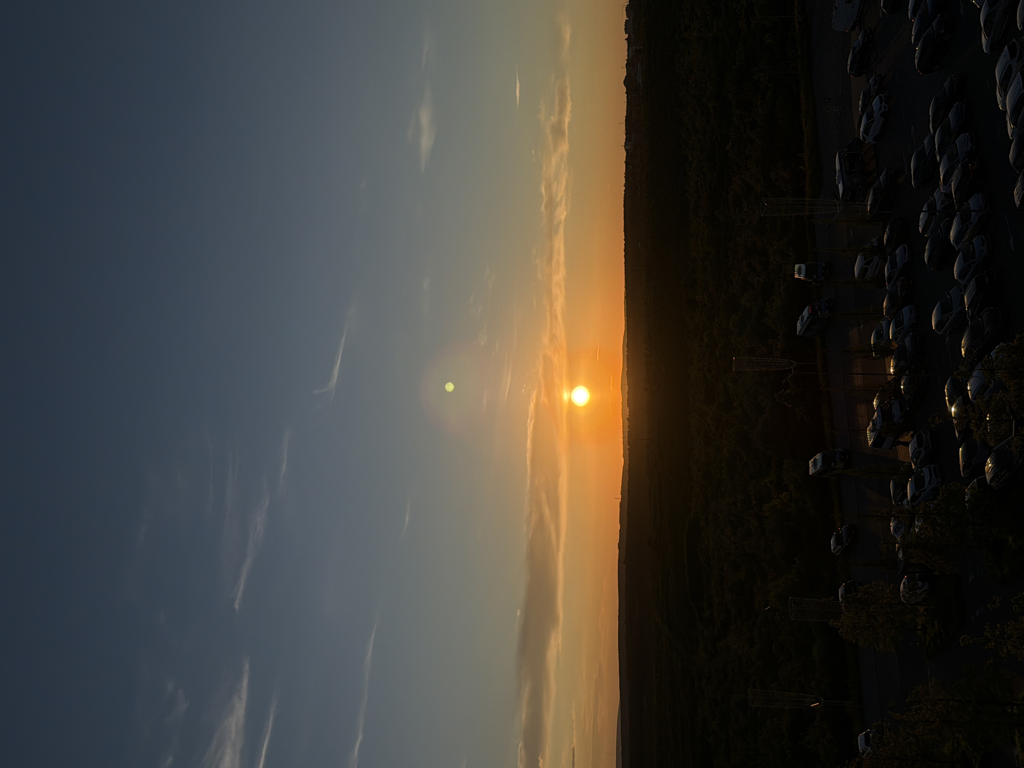 The image depicts a sunset over a sparsely populated area, likely the outskirts of Stăuceni, Moldova. The sky transitions from a deep blue at the top to vibrant orange and yellow hues near the horizon, with thin, wispy clouds scattered across. The sun is a bright, distinct orb low in the sky, casting a warm glow. In the foreground, a dark, dense forest or wooded area stretches across the mid-ground, with a faint, flat landscape visible beyond it. Below the tree line, a parking lot is visible, filled with numerous cars of various models and colors. Some streetlights or architectural features are also present in the lower section of the image, illuminated by the fading sunlight. The overall impression is one of a peaceful evening scene in a semi-rural or suburban setting.