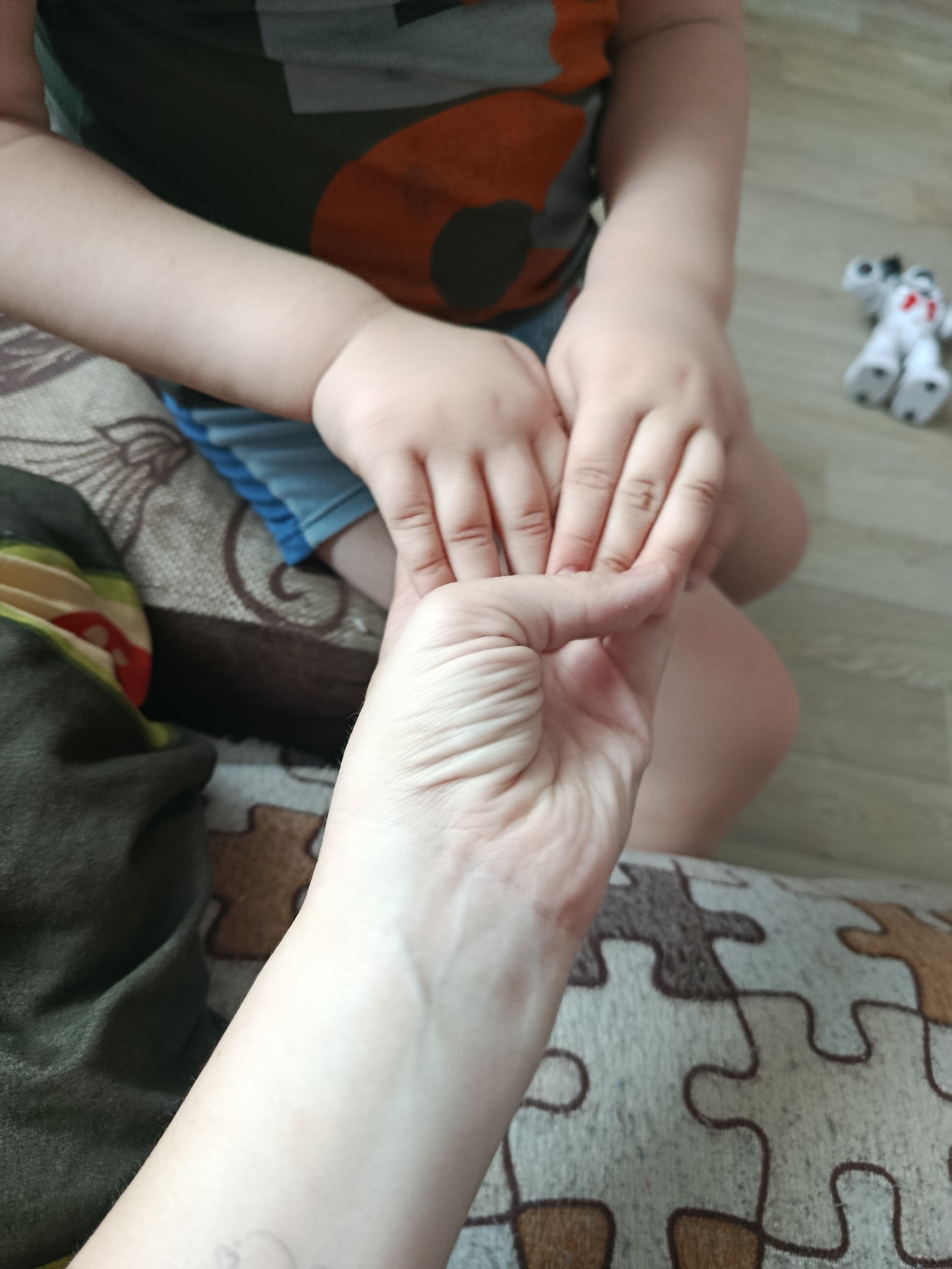 This close-up image depicts an intimate moment between an adult and a child in an indoor setting. The foreground features the lower arm and hand of an adult, characterized by visible wrinkles, gently holding the smaller, chubbier hand of a child. The child's other hand is also present, resting near the adult's hand.
The child is partially visible, wearing a dark green or grey t-shirt with an orange and black graphic design, and light blue shorts. They appear to be seated or kneeling on a surface covered with a patterned fabric, which has interlocking puzzle piece designs in shades of beige and brown. To the left, a dark green and striped fabric is also visible.
In the blurred background, a light-colored wooden floor is discernible, where a small, white and black toy, resembling a robot dog, lies. The lighting suggests it is daytime indoors. The scene conveys a sense of affection, comfort, and connection between the adult and child. There is no visible text within the image.