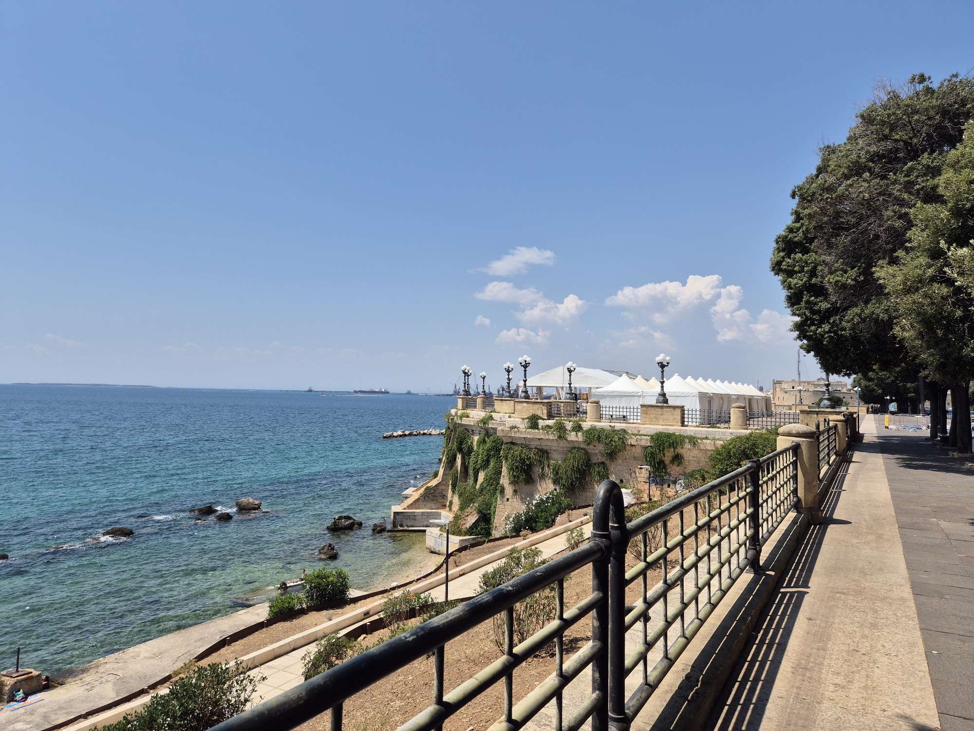 The image shows a scenic view of the coastline in Taranto, Italy. In the foreground, a black metal railing lines a paved walkway. The walkway is bordered by a dry, reddish-brown ground covered with sparse vegetation and a low stone wall. To the left of the walkway, the sea is visible, with clear turquoise water lapping against a rocky shore. Several large rocks are scattered in the shallow water. Above the shoreline, a stone embankment is covered with green foliage. On top of the embankment, there is a white-tented structure with white posts, and several lampposts with black tops. In the background, the sea stretches to the horizon, with a few ships visible in the distance. To the right of the walkway, a line of mature trees with dense green foliage casts long shadows on the pavement. The sky is a clear, bright blue with a few scattered white clouds. The overall impression is of a sunny day in a coastal city.