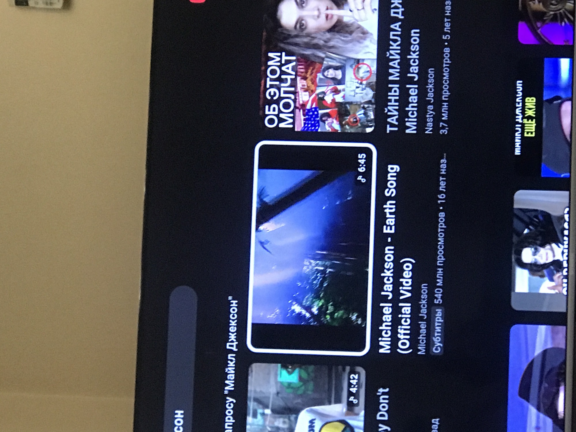 Here is a forensic description of the image:
The image appears to be a close-up of a television screen displaying the YouTube app. The screen shows search results related to Michael Jackson. Several video suggestions are visible, including "Michael Jackson - Earth Song (Official Video)" and a video with the title "Secrets of Michael Jackson".
There are no people present in the image, only thumbnails from the displayed videos. The environment consists of a room with an off-white wall and the television. The image does not contain enough information to determine the location in Yerevan, Armenia, or if it's even located in Armenia.