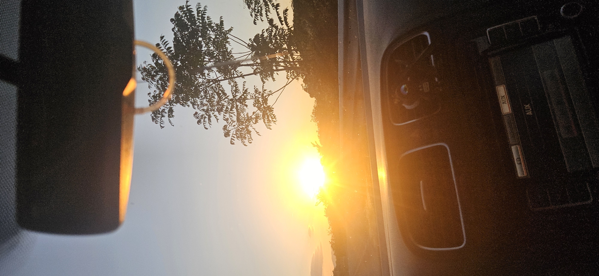 The image shows an interior car scene with a view through the windshield of a sunset and tree. The car's rearview mirror is visible on the left and the car's radio console is visible on the right. The radio has buttons labelled "AUX" and "CD". The location is potentially Mokrishte, Bulgaria, based on the prompt.