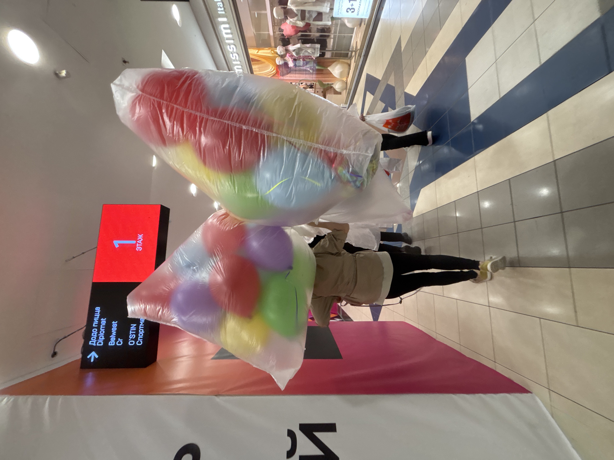 Two individuals, seen from behind, are walking down a tiled hallway. They are each carrying large, clear plastic bags filled with colorful balloons. The individual on the left is wearing a tan jacket and black pants. The individual on the right is wearing black pants and white shoes. In the background, a digital sign displays store directories and a floor number. A storefront with mannequins is also visible to the right. The environment appears to be an indoor shopping mall.