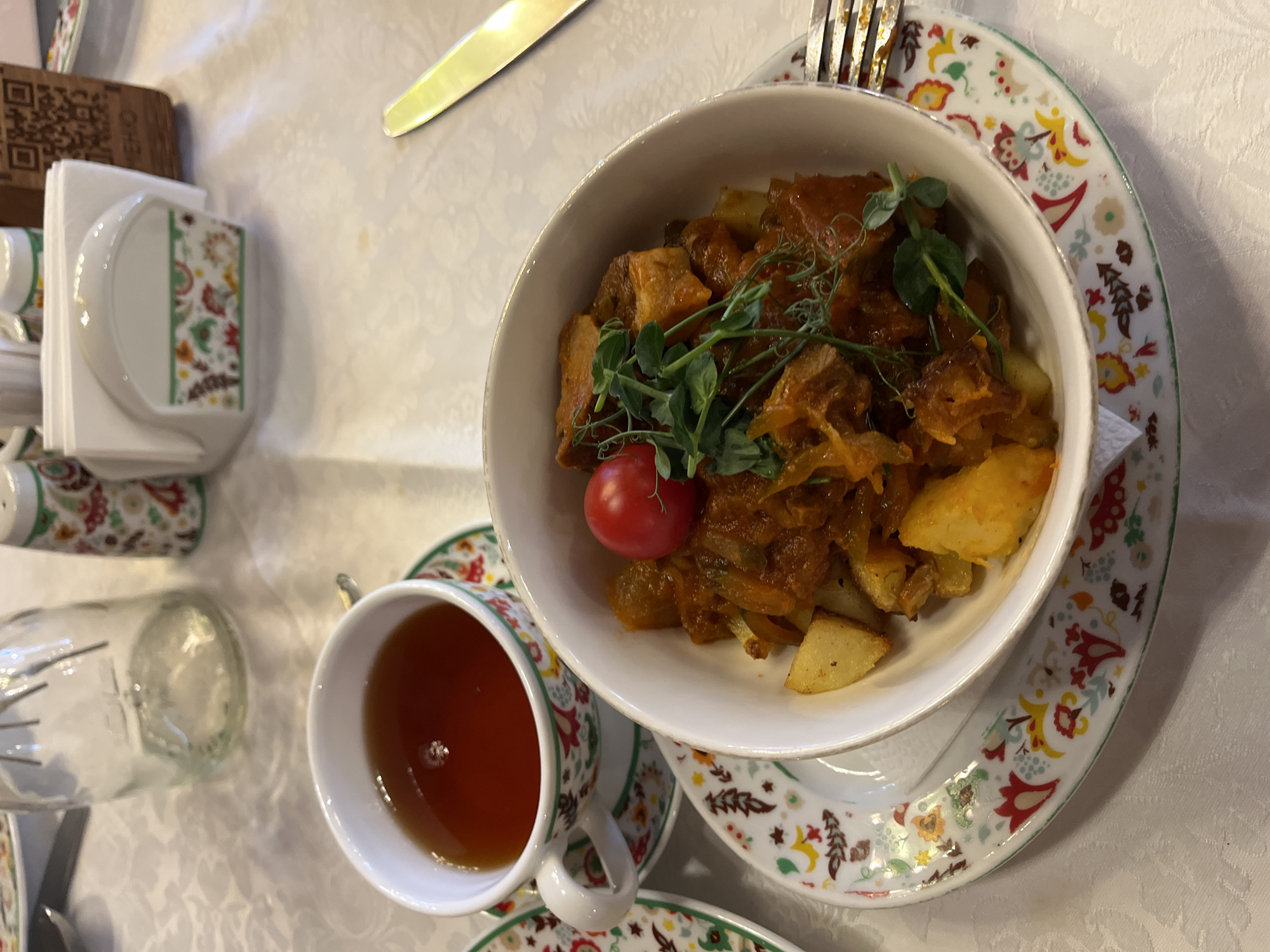 A table setting in Kazan, Russia, features a bowl of food, likely a stew or goulash with potatoes, garnished with sprouts and a cherry tomato. The bowl sits on a decorative plate. A cup of tea is visible beside it. A fork and knife rest on the table, along with a napkin holder.