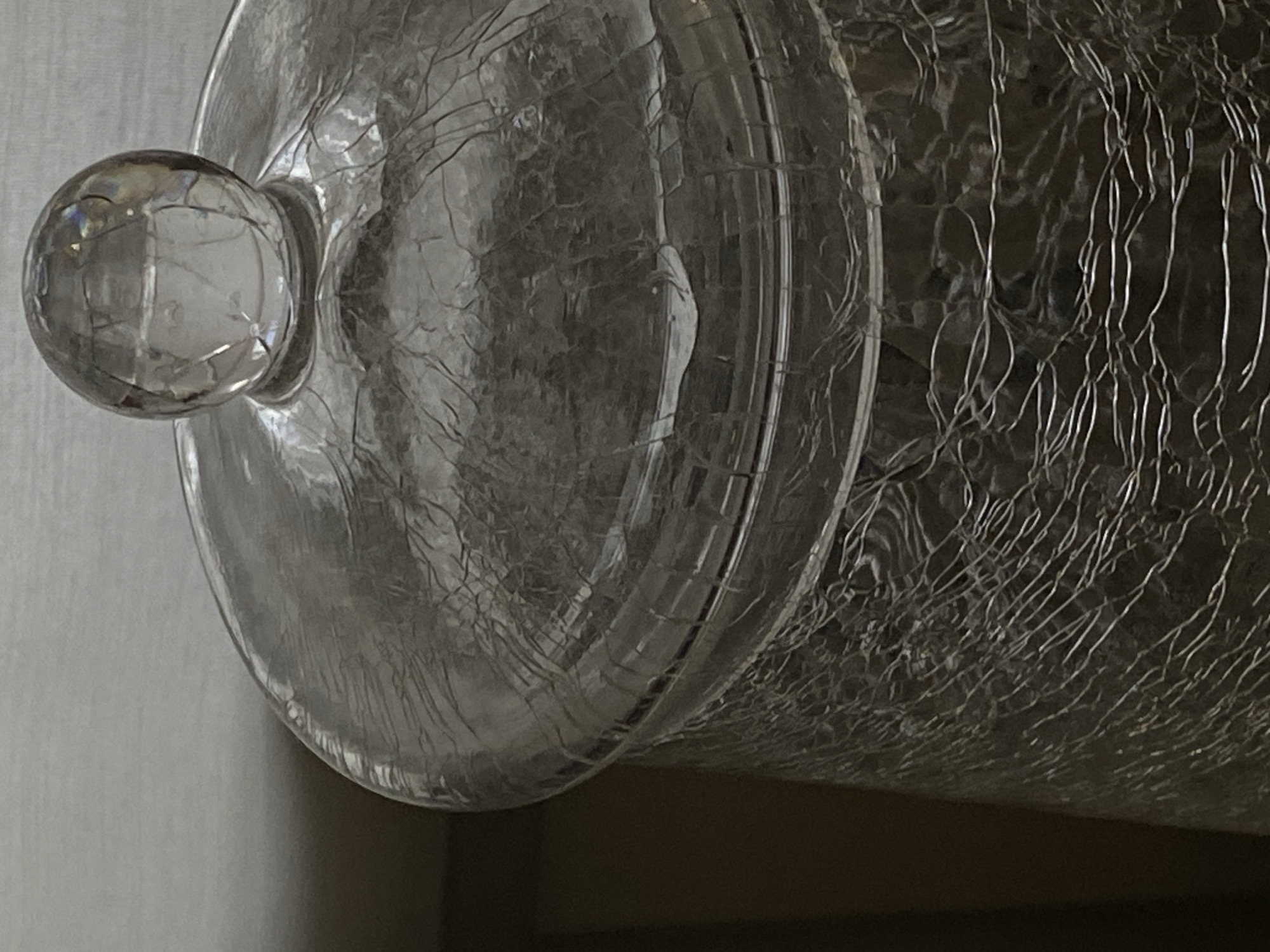 A clear glass jar with a decorative "crackle" texture is depicted in a close-up, slightly elevated shot. The jar features a circular lid topped with a spherical finial. The background consists of a plain, light-toned surface receding into an indistinct darker area on the left. No individuals, actions, or interactions are visible. The image contains no discernible information to specify a location within Votkinsk, Russia.