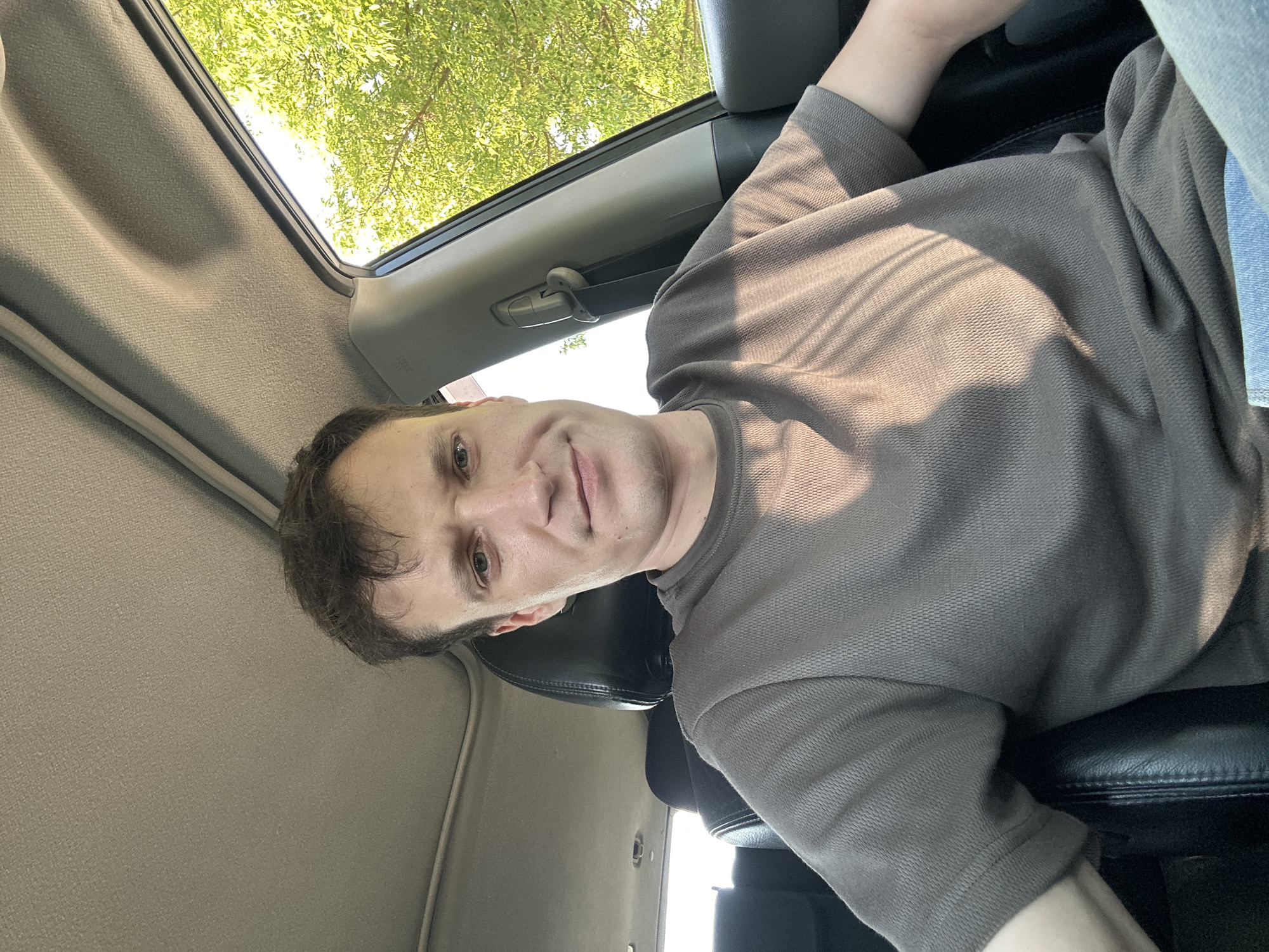 A man is sitting in the driver's seat of a car. He is wearing a brown t-shirt and blue jeans. He has short brown hair and is looking at the camera. The car is parked on a street in Makiv, Ukraine. There are trees and foliage visible outside the car window. The sun is shining and casting shadows inside the car.