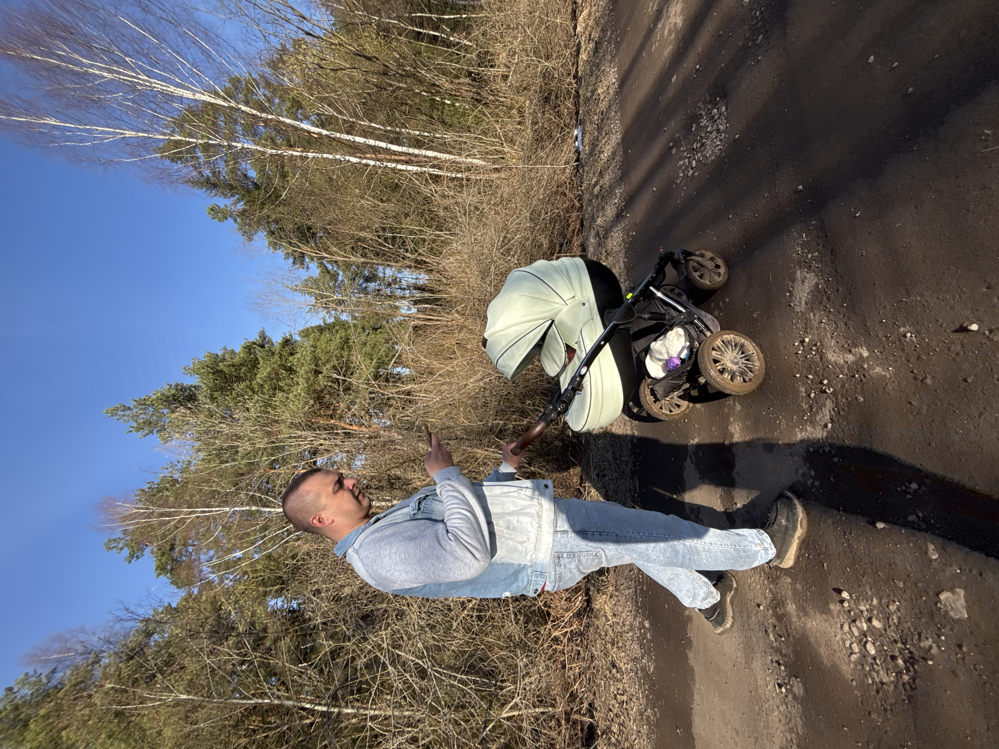 A man in a light-wash denim jacket and jeans stands on a paved road, pointing with his right hand towards a light green stroller. The road is adjacent to a wooded area with bare trees and some evergreen trees. The sky is blue. The location is Раздолье, Russia.