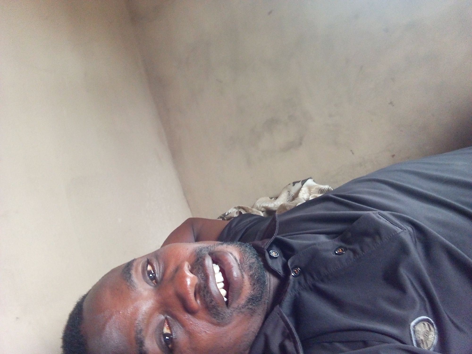 The image shows a dark-skinned man with a beard and short black hair. He is wearing a black button-down shirt and appears to be lying on his back, based on the angle and the wall visible in the background. His mouth is open, showing his teeth. The background is a plain, light-colored wall with some minor discoloration. There is no verifiable information that places this image in Adonkolo, Nigeria. The location is unknown.