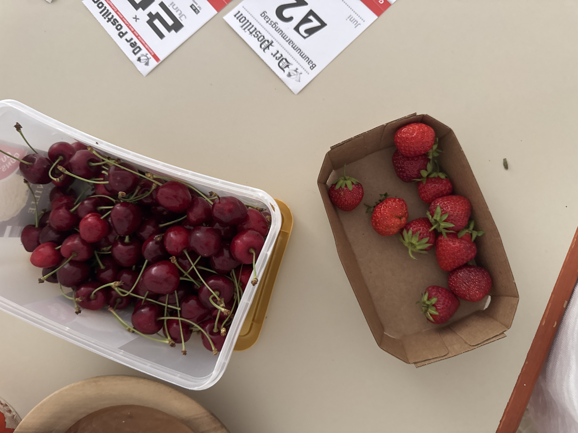 A top-down, close-up shot shows a container of cherries next to a small brown cardboard container filled with strawberries. The cherries are deep red and have green stems. They are in a clear plastic container with a yellow bottom. The strawberries are bright red and appear fresh, with green stems and leaves. Two small, white calendar pages are visible on the right side of the image. The background is a light beige surface, likely a table. The scene appears to be in Hohenbrunn, Germany, as indicated by the calendar pages which feature text such as "Der Postillon" and "Baumumarmungstag," common German calendar notations. There are no people or animals visible in the image, and no actions or interactions are taking place. The lighting is natural, casting soft shadows.