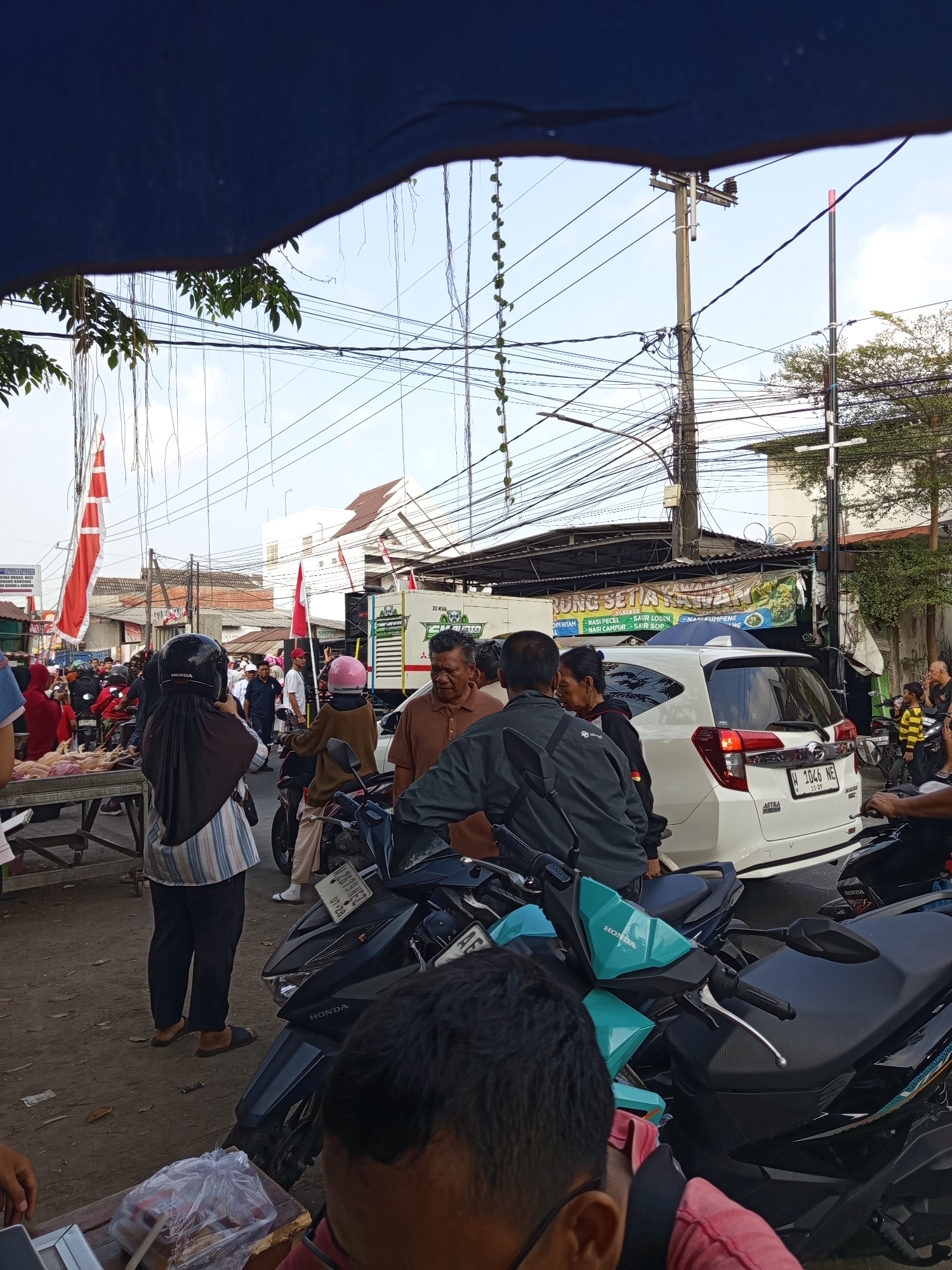 This is a street scene in Suko, Indonesia, likely a market, with a crowd of people, many of whom are on motorcycles. The foreground shows several motorcycles parked closely together, with their handlebars and seats visible. A person in a red shirt is partially visible in the bottom right corner, and another person wearing a mask and glasses is in the bottom center.
In the mid-ground, a white SUV is parked, and several people are standing around it. To the left of the SUV, there is a market stall with raw chickens displayed. A woman in a hijab and striped shirt is standing near the stall. People are also seen on motorcycles in this area. Red and white flags, possibly national flags, are visible in the background, along with power lines and buildings.
The environment appears to be a busy street with a mix of residential and commercial buildings. The sky is overcast, suggesting it might be morning or late afternoon. The overall impression is a vibrant, active street scene typical of a local market in Indonesia.