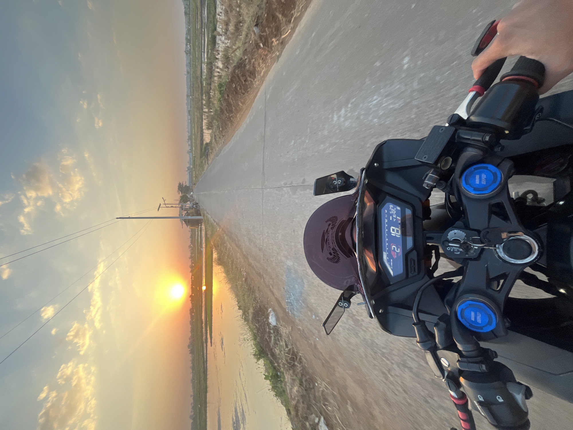 The image captures a first-person perspective of riding a motorcycle along a rural road in Pecangaan, Indonesia, during sunset. The sun is low on the horizon, casting a warm golden glow across the sky and reflecting on a body of water to the left of the road. The road is asphalt and stretches into the distance, flanked by fields of rice paddies on the right and the water on the left. Power lines are visible overhead. The motorcycle's handlebars and dashboard are in the foreground, with a rider's hands gripping the handlebars. The dashboard displays the gear as "2" and the time as "5:29". Small buildings and structures are sparsely scattered in the background along the road.