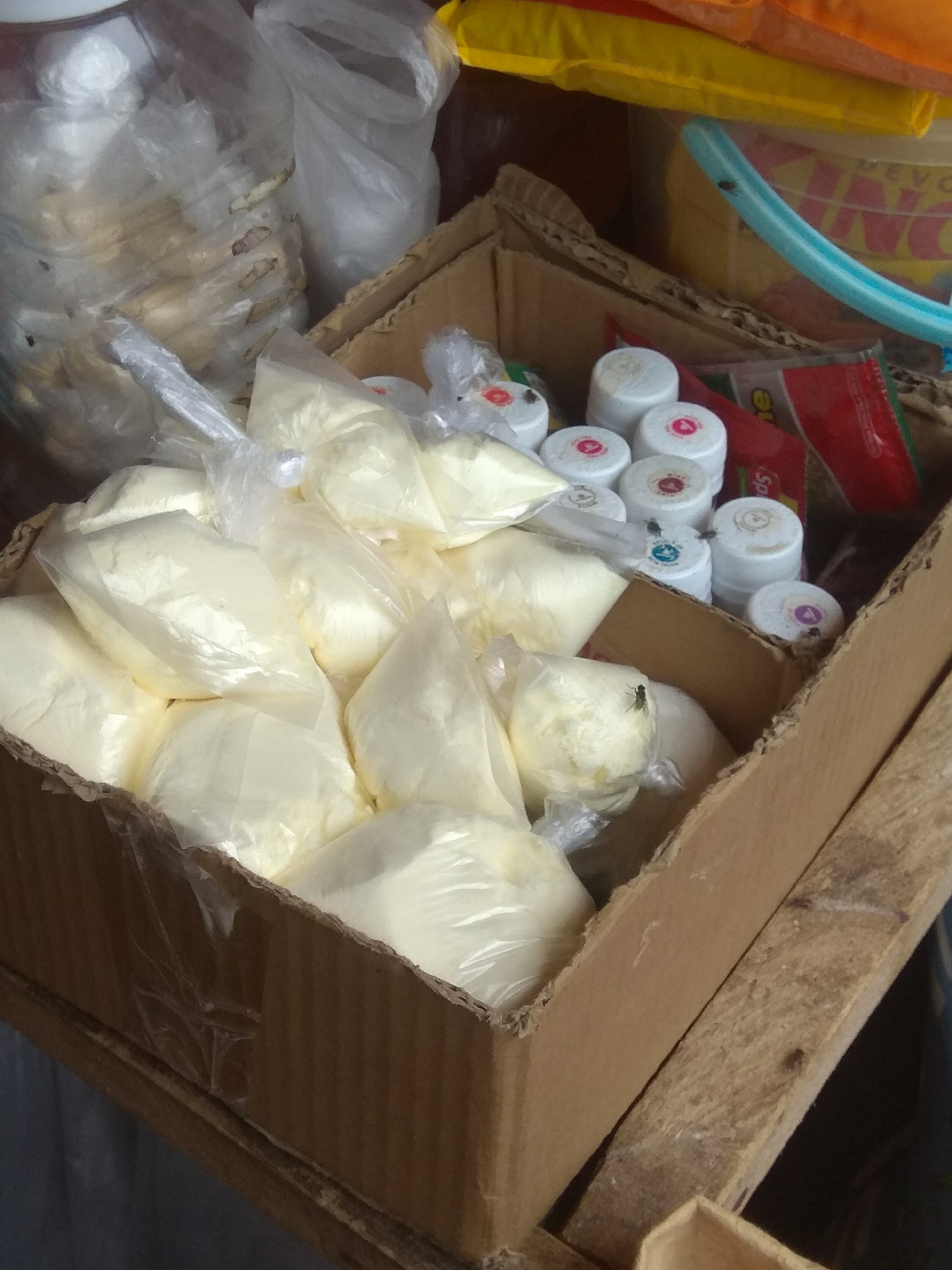 The image depicts a close-up view of a cardboard box filled with numerous individually bagged, pale yellow powdery substances. The bags are transparent plastic, allowing the contents to be visible. Several small, white containers with colored tops are also present within the box, some with visible labels. A few flies are present, resting on some of the containers and bags. The box itself appears to be made of corrugated cardboard and is partially filled. The background is blurred but suggests an indoor environment, possibly a market stall or storage area. The scene appears to be located in the city of Jalingo, Nigeria, although no specific landmarks or identifying features confirm this. The overall impression is one of storage or preparation of ingredients or products.