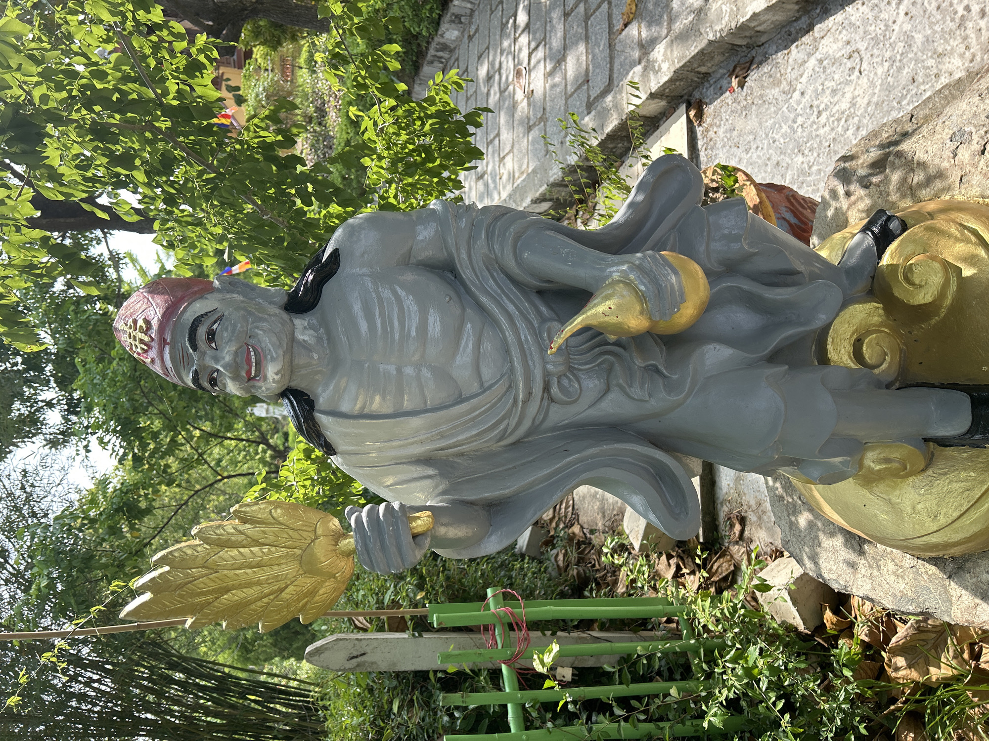 A weathered, grey statue of a Buddhist deity stands outdoors in what appears to be a temple or garden setting in Nam Nha Trang Ward, Vietnam. The statue, depicted with a jovial expression, wears a red cap adorned with a gold symbol and holds a golden fan-like object in its right hand and a golden gourd in its left. It is positioned on a golden base resembling a cloud. The background features lush green foliage, bamboo stalks, a stone pathway, and hints of temple architecture in the distance. The lighting suggests a sunny day.