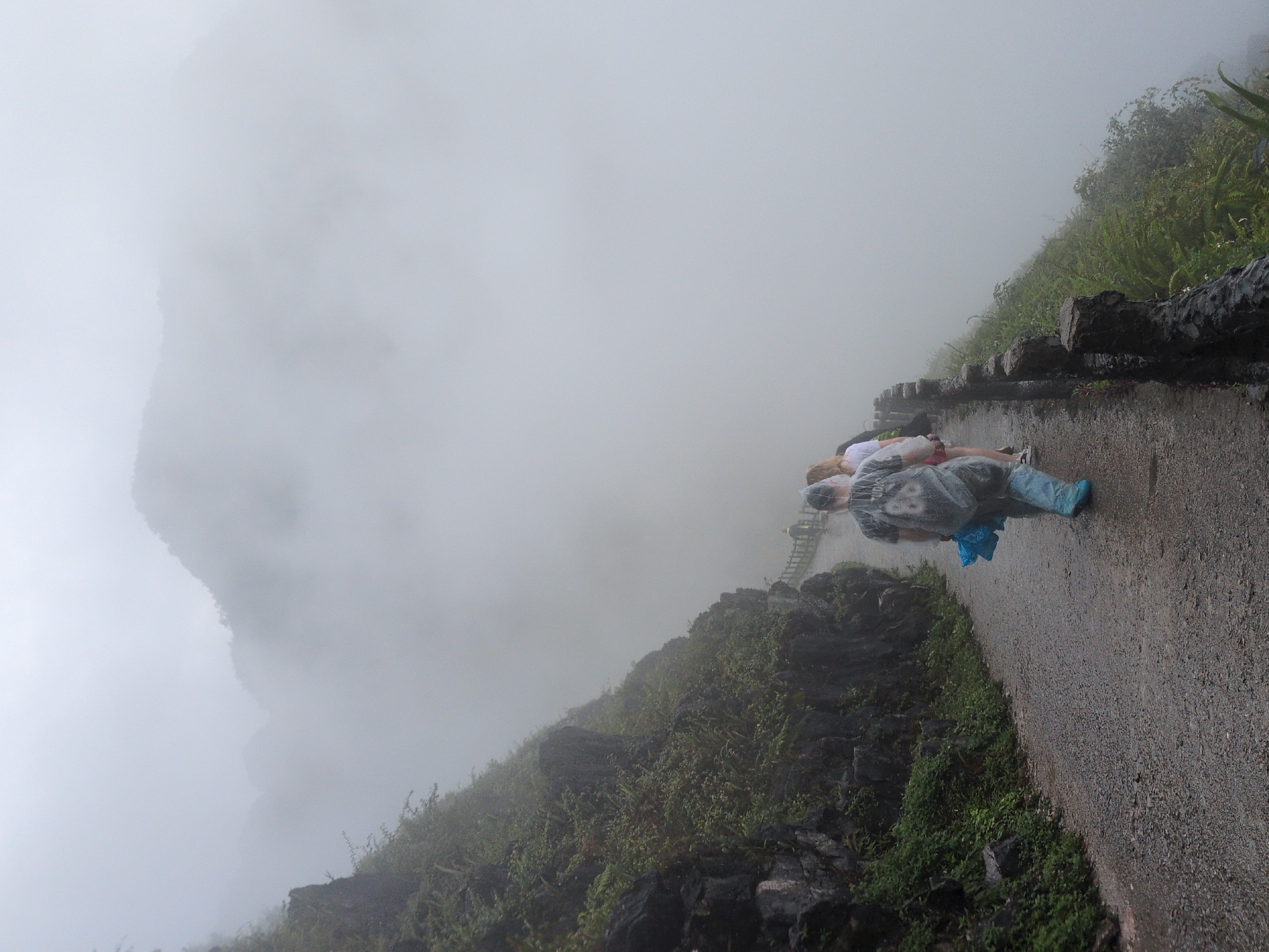 A paved pathway winds along a steep, vegetated cliffside under a heavy, foggy sky. Several people are walking along the path, some wearing ponchos. The fog obscures the distant mountains and creates a sense of depth and mystery in the landscape. The pathway itself appears to be made of gravel and is wet, suggesting it might be raining or has recently rained. A wooden railing lines the edge of the path on the side that drops off into the fog. The vegetation on the cliffside is lush and green, with many small trees and plants clinging to the rocky surface. The overall atmosphere is moody and dramatic, with the fog playing a significant role in shaping the visual experience. This scene is characteristic of the dramatic karst landscapes found in Đồng Văn Commune, Vietnam, particularly at viewpoints overlooking the valleys and mountains.