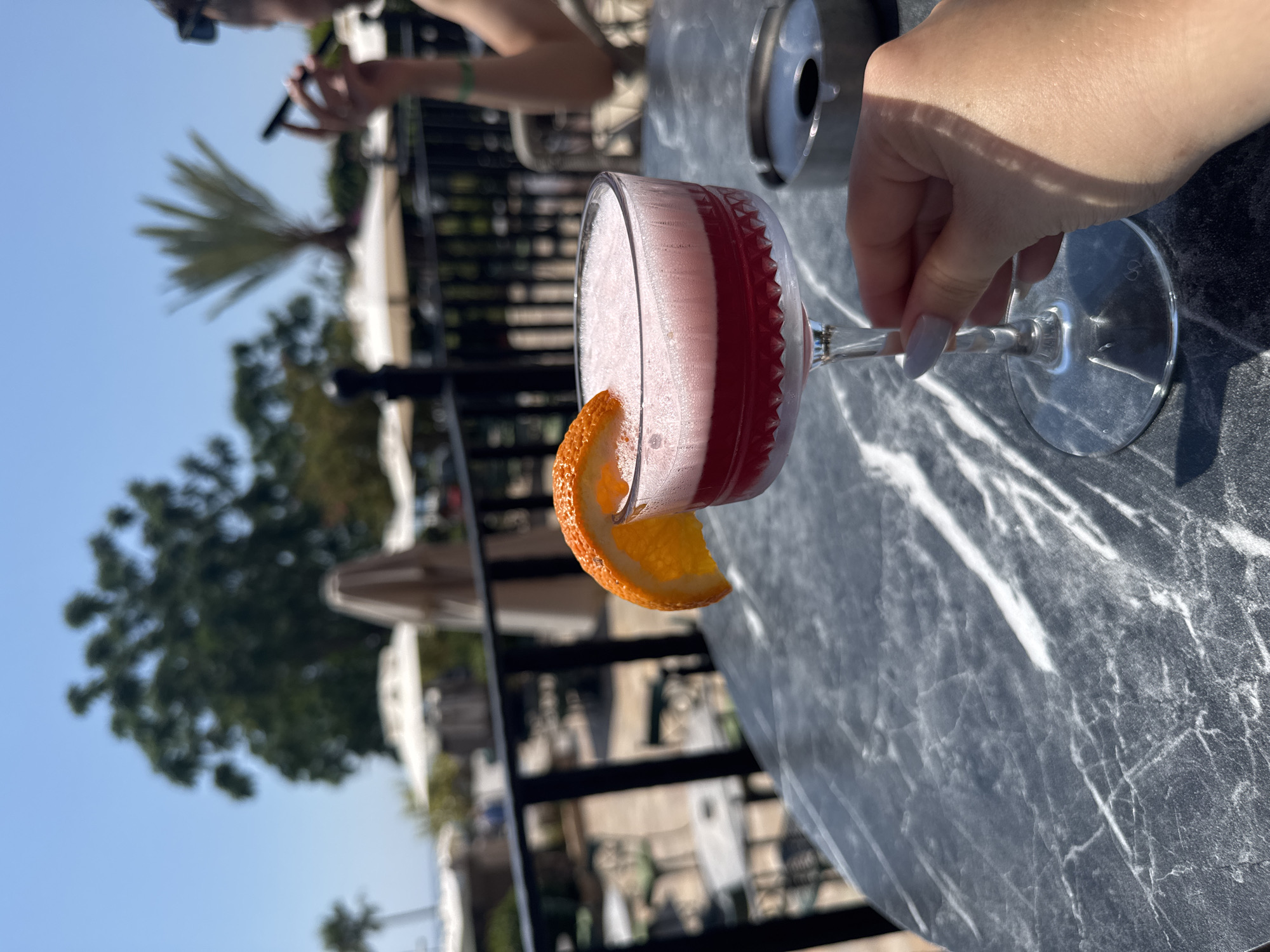 A woman's hand with light purple nail polish is holding the stem of a cocktail glass filled with a pink foamy drink topped with an orange slice. The glass is placed on a dark marble table. An ashtray is visible to the right of the glass. In the background, there is an outdoor seating area with chairs and tables, umbrellas, and palm trees. The scene appears to be at a resort or restaurant in Kemer, Turkey, given the clear blue sky and Mediterranean vegetation. A person is partially visible in the far background, holding a phone.
