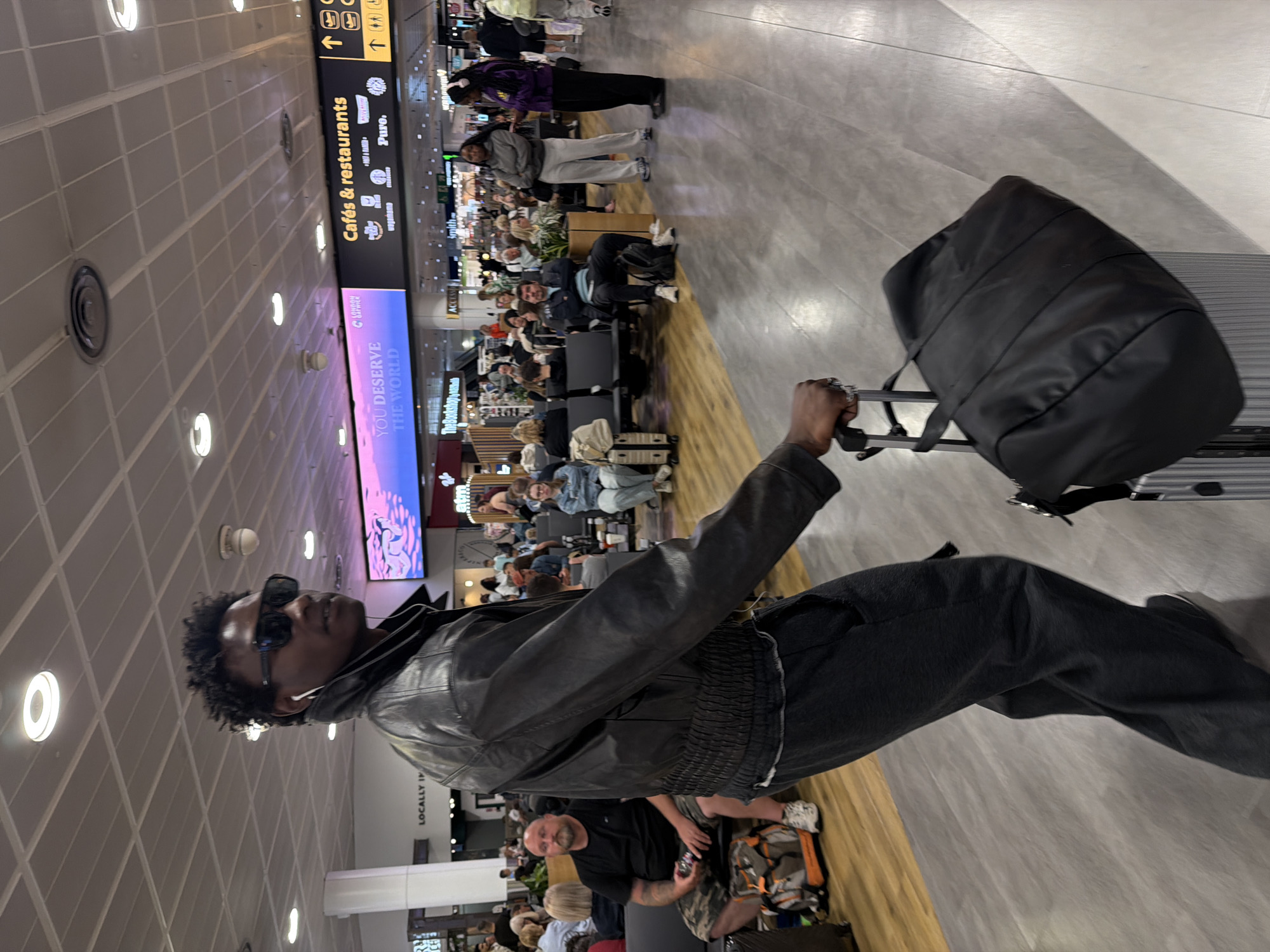 The image depicts a young Black male walking through an airport terminal, likely Gatwick Airport, Crawley, United Kingdom, based on the overhead signage for "Cafés & restaurants" and brands like "Wagamama." He is wearing dark sunglasses, a black leather jacket over a grey t-shirt, and dark sweatpants. He has white earbuds in his ears and is pulling a large black duffel bag on top of a silver rolling suitcase with his right hand.
The terminal floor is a light grey tile, with a wooden section on the left where other travelers are seated on grey benches. In the background, there are advertisements on large screens, including one that reads "YOU DESERVE THE WORLD." People are seated in waiting areas, and others are walking in the distance. The ceiling is a grid of white panels with circular light fixtures. The overall environment suggests a busy airport departure lounge.