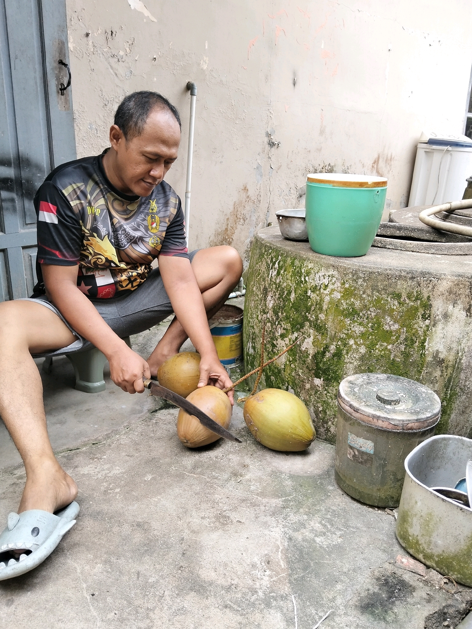 A man is crouched on the ground in front of a weathered, off-white wall and a concrete surface with scattered debris. He is wearing a black t-shirt with a multi-colored graphic design and grey shorts. His left leg is bent, and his right leg is extended forward. He is holding a large knife in his right hand and is in the process of cutting a coconut. Three coconuts are positioned on the ground in front of him; one is partially cut. Next to the coconuts, to the man's right, is a large, circular concrete structure covered in green moss. On top of this structure are a small silver bowl and a green bucket with a wooden rim. To the right of the concrete structure are two cylindrical containers, one dark green and one silver, and a white bucket containing items. A grey slipper shaped like a shark is on the man's left foot. The setting appears to be an outdoor area, possibly a backyard or alleyway, in Arang Limbung, Indonesia.