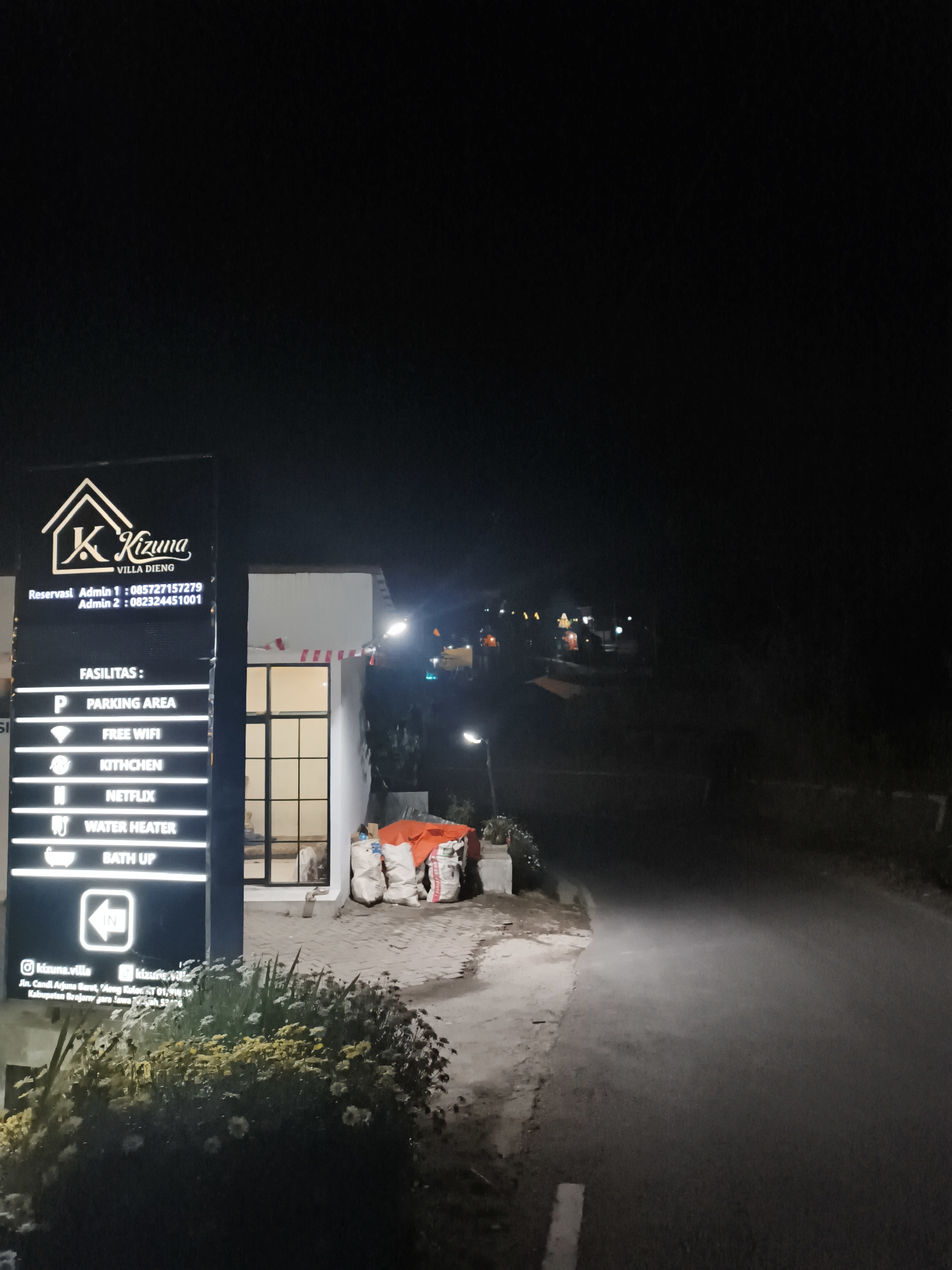 A vertical sign for "K Kizuna VILLA DIENG" stands illuminated at night in front of a building. The sign lists "FASILITAS" including Parking Area, Free WiFi, Kitchen, Netflix, Water Heater, and Bath Up, along with icons. Reservation contact numbers are also listed. Below this, social media handles "@kizuna.villa" and "@kizuna.villa" are visible, along with the address "Jln. Candi Arjuna Barat, 'Aeng Kulon, RT 01, RW 12, Kabupaten Banjarnegara." In the background, a dimly lit building with windows is visible, and piles of bagged materials, including orange tarps, are stacked near the entrance. The area appears to be a road with some vegetation, including a bush with yellow and white flowers in the foreground. The scene suggests a commercial establishment, likely a villa or guesthouse, in a rural or semi-rural setting.