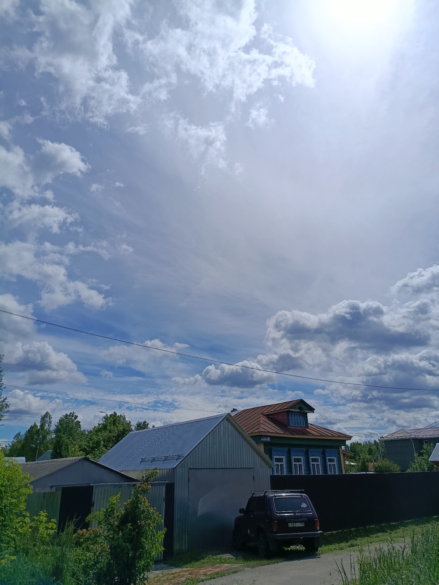 The image depicts a scene in the city of Vereya, Russia, under a partly cloudy sky. In the foreground, a gravel driveway leads past lush green grass and foliage. To the left, a dark-colored SUV is parked next to a metal garage with an open door. Behind the SUV and garage, a traditional Russian wooden house with a red roof, green siding, and blue-trimmed windows is visible. A dark metal fence runs behind the house. Further in the background, more trees and buildings can be seen, along with utility lines stretched across the sky. The sun is bright, casting a glare in the upper right portion of the image.