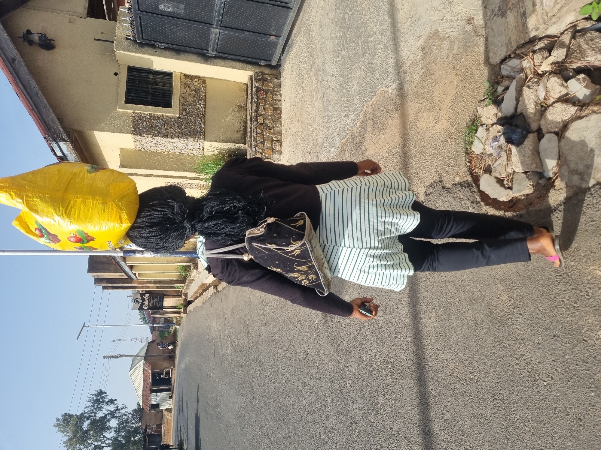 A woman walks on a paved street in Jos, Nigeria, carrying a large yellow bag on her head. She is dressed in a black top, a striped skirt, and black pants, and wears sandals. A patterned bag hangs on her shoulder. She is holding a mobile phone in her right hand. The background shows buildings, utility poles, and trees under a clear sky. A building stands to the right with a gray metal door and a window with bars. A rock border runs alongside the street.