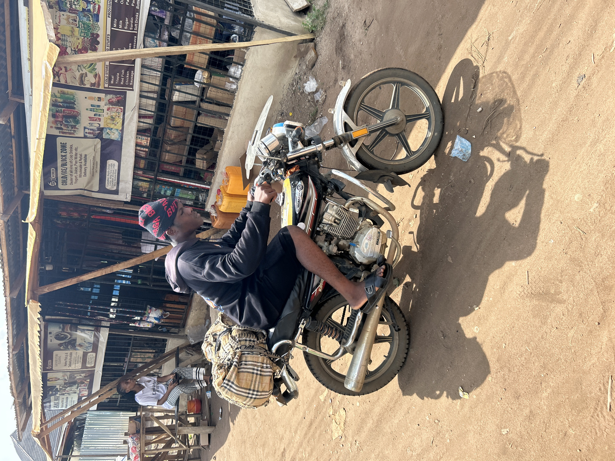 A man is sitting on a parked motorcycle in Ihiagwa, Nigeria, next to a roadside shop. The shop displays signs advertising "Cold/Ice/Block Zone" drinks, laundry services, and food items like meat, fish, milk, and soft drinks. Another person sits near the shop on a wooden structure. The ground is dirt.