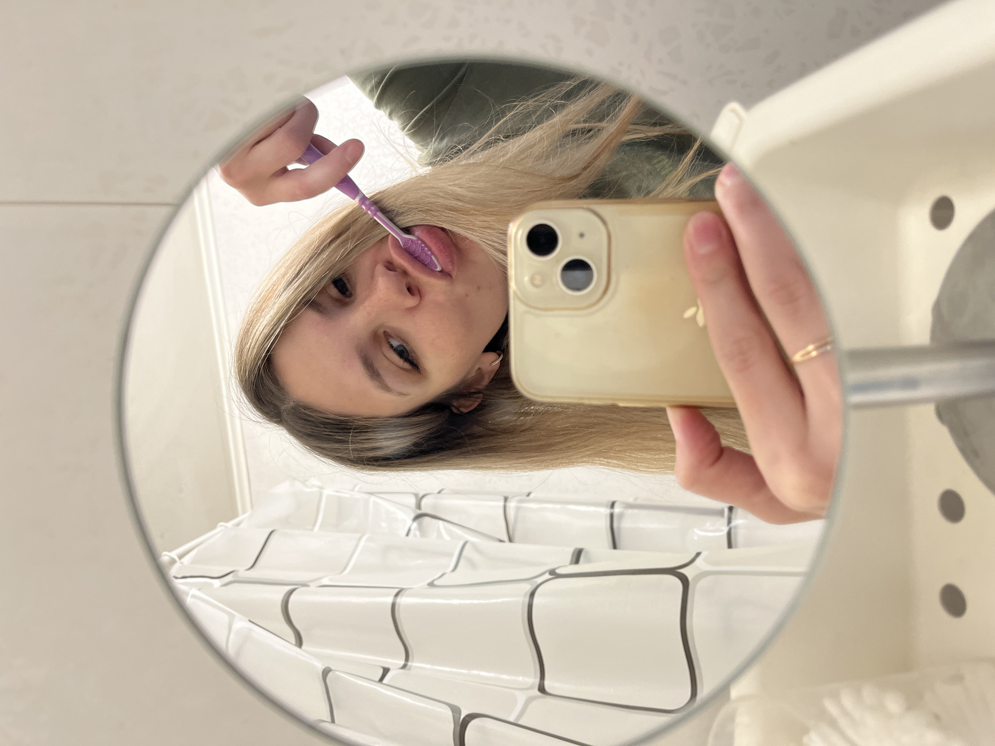 A woman is visible in a round mirror. She is holding a gold-colored smartphone in her right hand, presumably to take the picture, and brushing her teeth with a purple toothbrush in her left hand. The reflection reveals she is in a bathroom. A white sink and a white shower curtain with a gray-bordered tile pattern are also visible. There is no definitive evidence to confirm the location is Chaikovsky, Russia.