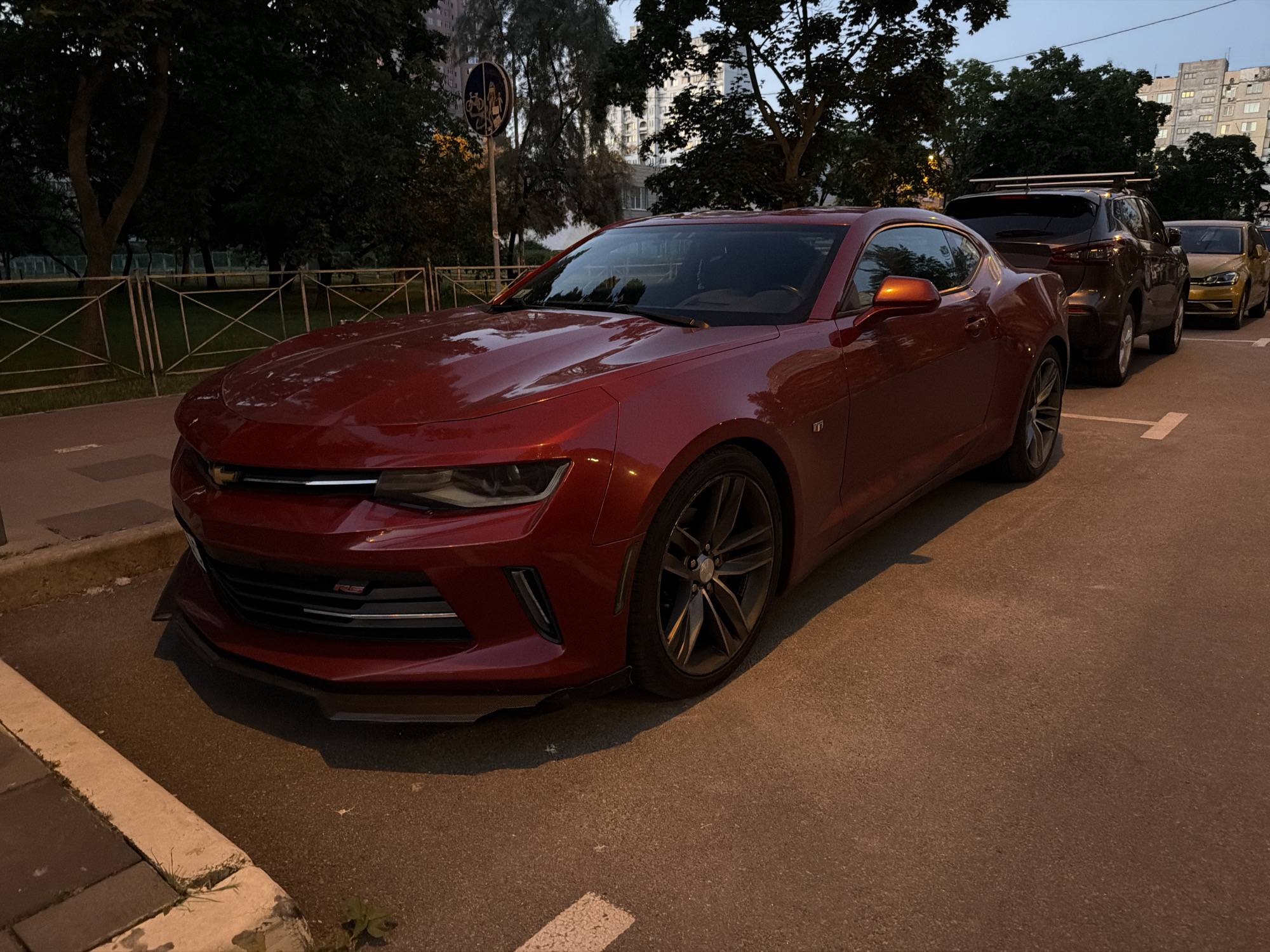 In a city environment, a red Chevrolet Camaro is parked on an asphalt road next to a sidewalk. The car is the primary subject, positioned in the foreground and taking up a significant portion of the frame. Its front end, particularly the grille and headlights, are visible, as is a portion of its side profile and a prominent wheel. The car appears to be clean and in good condition.
Behind the Camaro, a dark-colored SUV is parked, followed by a light-colored car, suggesting a parking area or street. The background includes trees with dark foliage, a metal fence, and in the distance, buildings that appear to be residential. A street sign with pictograms for a bicycle and a pedestrian is also visible on a pole. The lighting suggests it is either early morning or late evening, as the sky is a muted blue and there are shadows cast by the trees and vehicles. The ground appears slightly damp, possibly from recent rain.