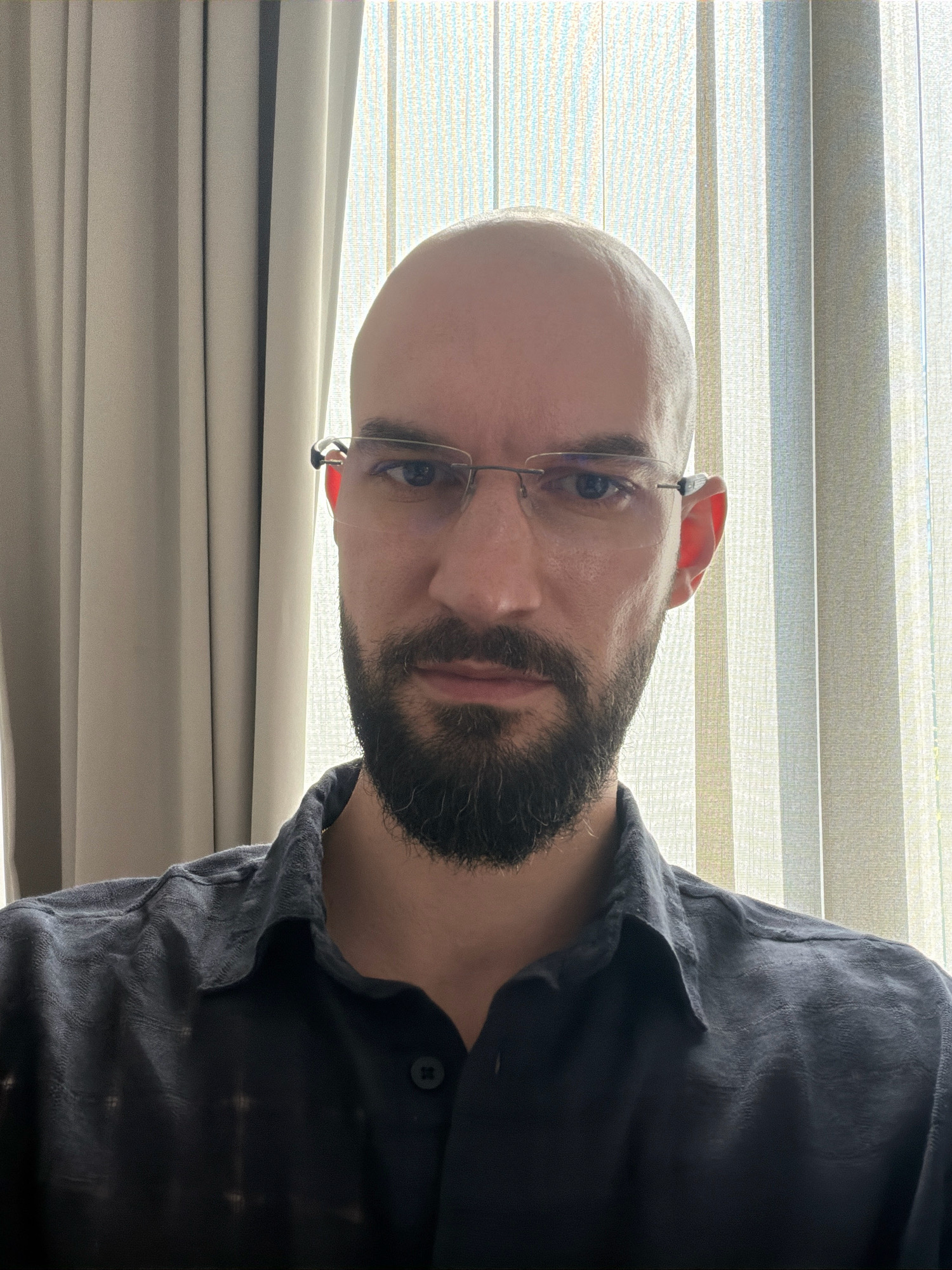 The image is a close-up selfie of a bald, Caucasian man with a beard and glasses. He is wearing a dark, collared shirt. He is positioned in front of vertical blinds, with a curtain visible on the left side of the frame. The lighting suggests an indoor environment. There is no indication of the specific location in Sofia, Bulgaria.