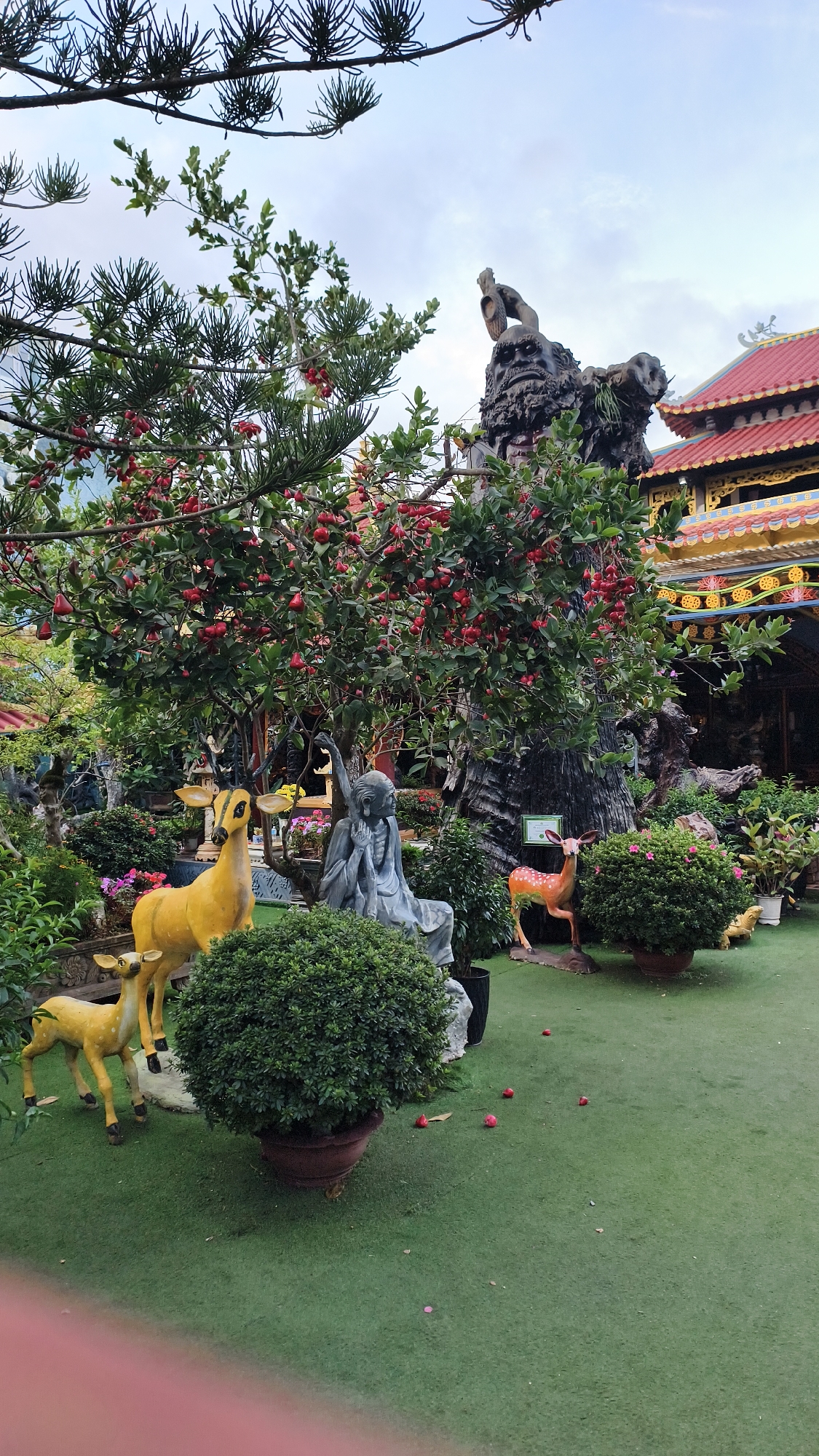 The image depicts an outdoor courtyard in Xuân Thành, Vietnam, characterized by a garden setting with artificial elements and foliage. In the foreground, a bright green artificial turf covers the ground. To the left, two yellow statues of deer are visible, one larger and one smaller, positioned on a raised platform with a decorative border. Next to the deer, a gray statue of a meditating figure, possibly a Buddha or a monk, is seated. In the center of the image, a large tree laden with red fruits is prominent. Behind the tree, a dark, textured sculpture or natural formation rises, topped with a figure resembling a weathered face. To the right of the central tree, another statue of a deer, this one orange and white, stands. The background features a building with a red tiled roof and ornate yellow trim, suggesting a temple or a traditional Vietnamese structure. Several potted plants, including a large, round bush in the foreground, are strategically placed throughout the courtyard. The sky above is overcast, with a few branches of a dark green, spiky tree extending from the top of the frame. Scattered red fruits lie on the green ground.