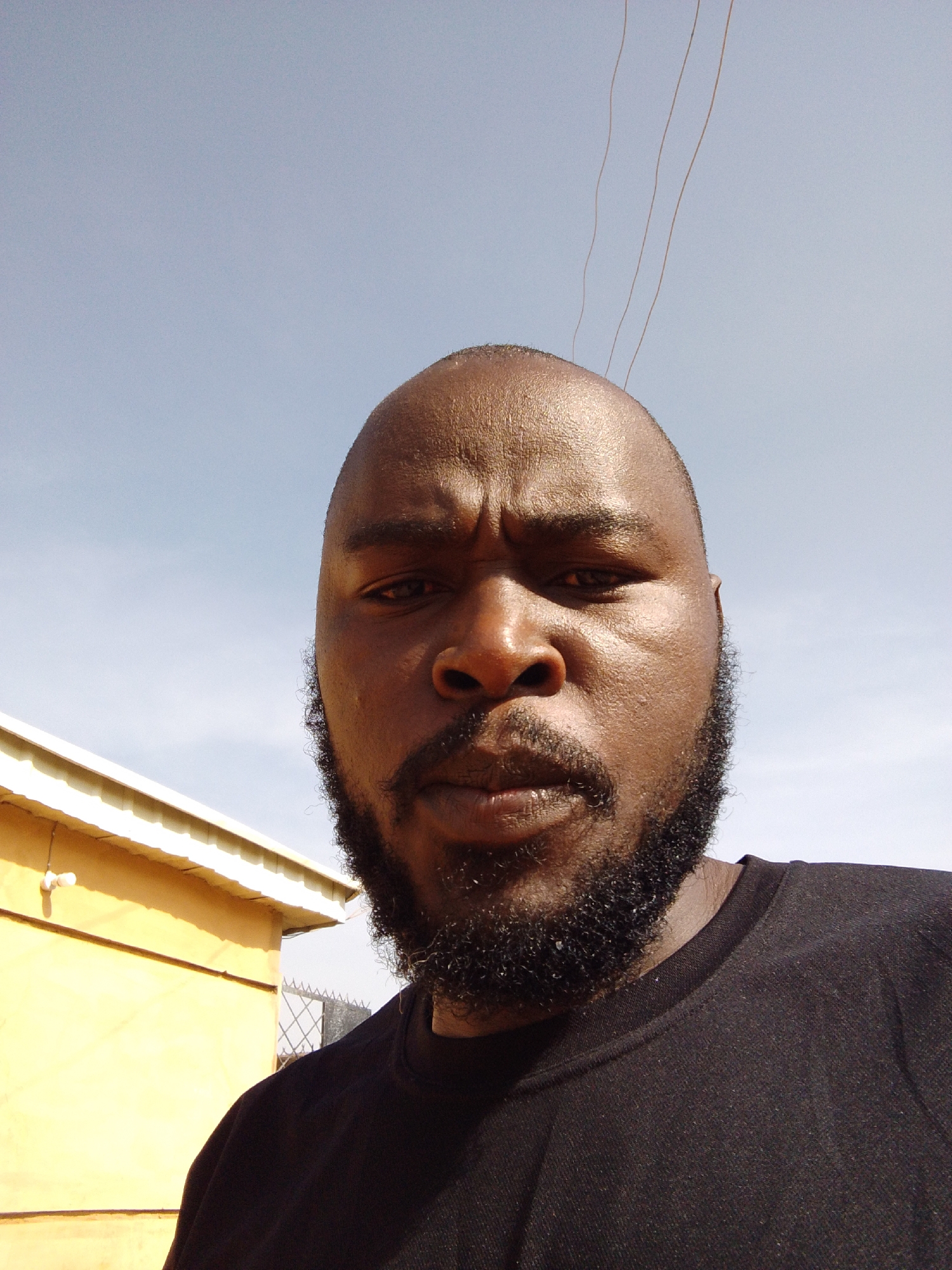 A close-up, low-angle shot of a dark-skinned man with a bald head and a full beard. He has a serious expression, his eyebrows furrowed. He is wearing a black t-shirt. Behind him and slightly to the left is a yellow building with a white roof. Above him is a light blue sky with some thin strands resembling wires. The photo may be taken in Jos, Nigeria.