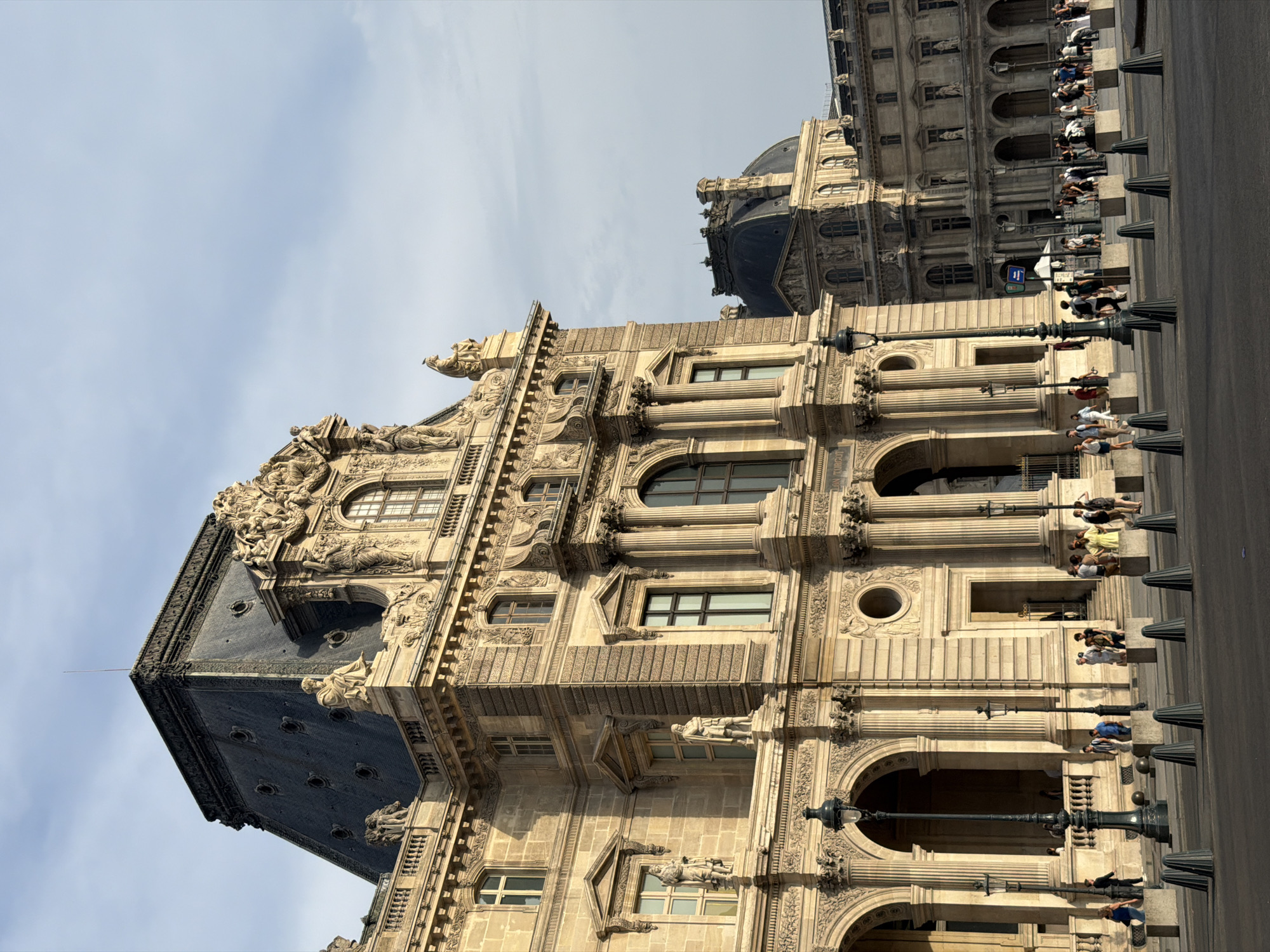 The image shows a wide-angle view of the Louvre Museum in Paris, France, on a clear day with some clouds. The architecture is ornate and classical, with large stone buildings featuring many windows, columns, and decorative sculptures. The main building in the foreground is a light-colored stone structure with a dark, mansard-style roof topped with intricate ornamentation. A statue of a seated figure is visible on the roofline.
In front of the building, there is a paved area where a group of people are walking and standing. They are dressed in casual summer clothing. Several lampposts with ornate designs are positioned along the walkway. Concrete bollards are arranged in rows on the paved area, separating it from the street. In the background, another section of the Louvre, featuring a dome, is visible. The lighting suggests it is either early morning or late afternoon, casting long shadows.