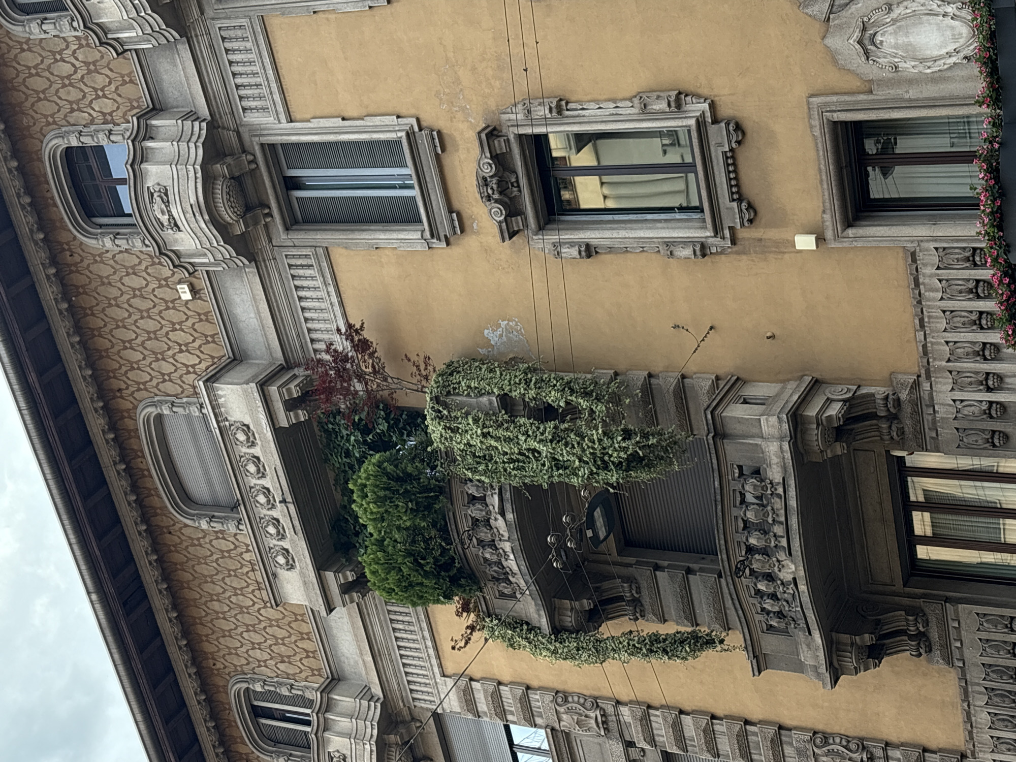 The image depicts a building facade in Milan, Italy. The facade is predominantly yellow with ornate architectural details in gray stone. There are several windows, some with blinds or shutters. A balcony with a railing is adorned with greenery, including vines and shrubs. There are electrical wires visible against the yellow wall. The sky is overcast.
