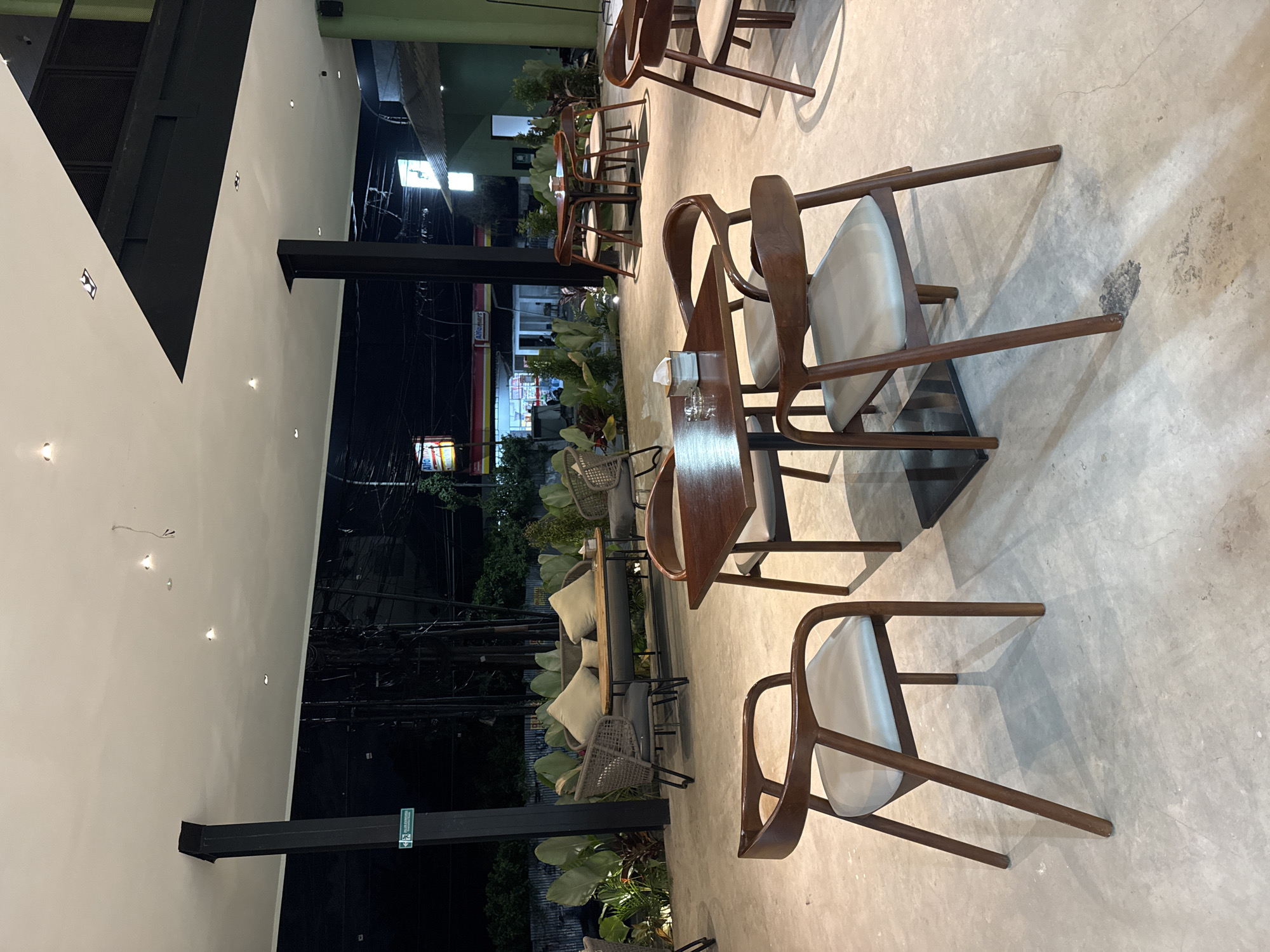 The image depicts an outdoor seating area of a restaurant or cafe, likely in Kuta Selatan, Indonesia. Several wooden chairs with light-colored cushions are arranged around small, dark-topped tables. The seating extends into an outdoor area where rattan-style furniture and cushions are visible. Lush greenery surrounds the seating area. In the background, the street is visible, illuminated by streetlights and shop signs. The image seems to have been taken at night.