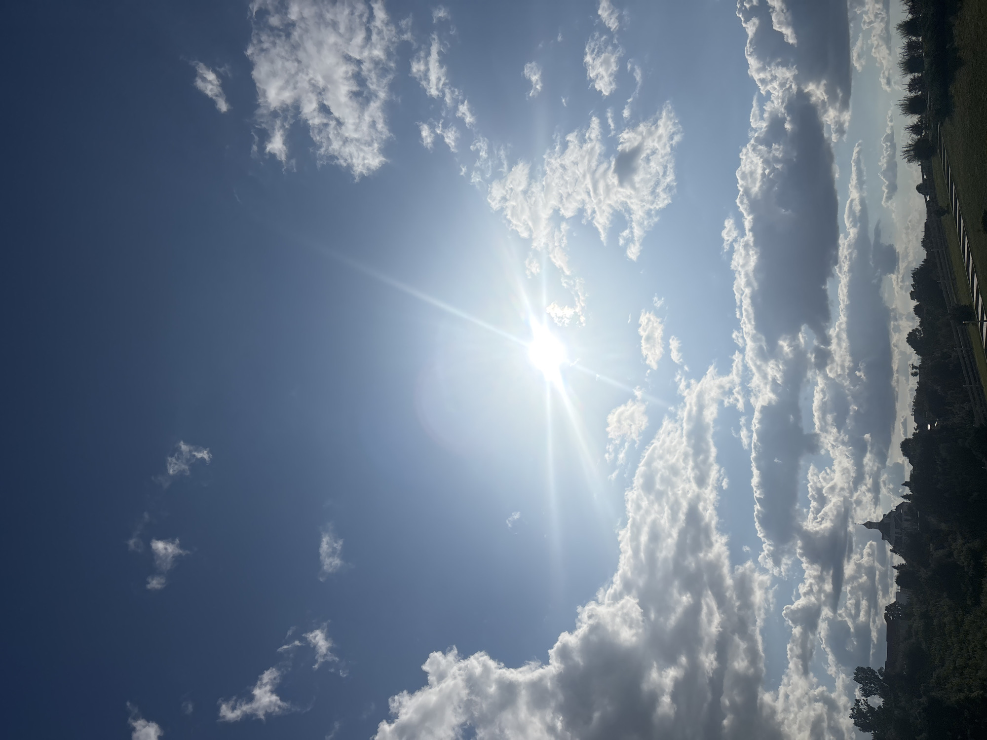 The image depicts a bright, sunny sky with scattered white and grey clouds, creating a dynamic and somewhat dramatic visual effect. The sun is prominently featured, emitting strong rays that suggest it is likely midday. The blue of the sky is vivid, indicating clear weather despite the presence of clouds.
To the right side of the frame, the scene transitions to a landscape. Dark green trees form a dense treeline, and beyond them, a building with a distinctive roofline and what appears to be a spire is visible. This structure is silhouetted against the bright sky, adding a point of interest to the composition. The ground below the trees is covered in lush green grass, suggesting a natural or park-like setting. A dark, linear element, possibly a fence or a path, runs diagonally from the bottom right corner towards the center of the image, separating the grassy area from the treeline.
The image's orientation is sideways, with the landscape elements appearing on the right side and the sky occupying the majority of the frame. The overall impression is one of a beautiful, clear day, with the sun's intense light filtering through the clouds and illuminating the landscape. There is no visible text in the image.