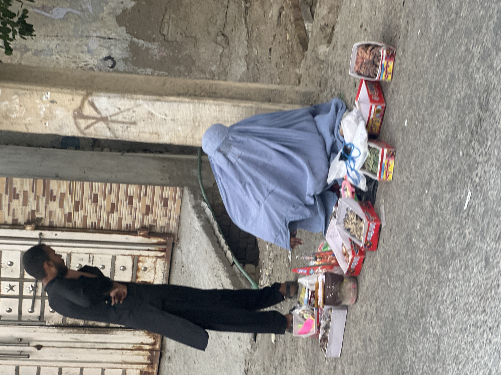 The image shows a street scene in Quetta City Tehsil, Pakistan. A woman in a blue burqa sits on the ground next to a display of boxes of what appear to be hair accessories or jewelry for sale on the pavement. A man in a black salwar kameez stands nearby with his arms crossed. The background includes a building with a tiled facade and a concrete sidewalk. The overall impression is of a low-income commercial area.
