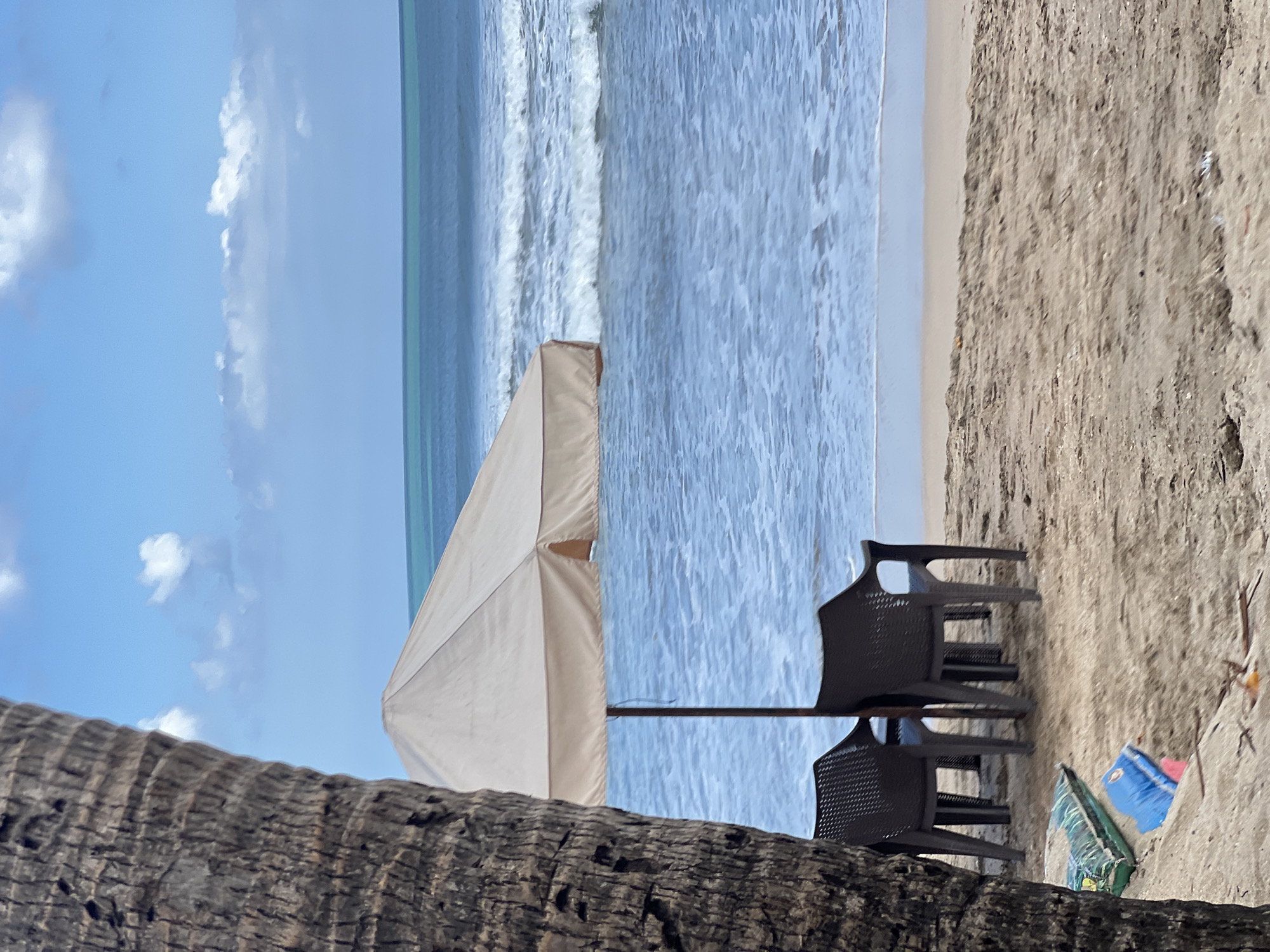 A beach scene in Kuta, Indonesia, features a palm tree trunk in the foreground, partially obscuring a beach umbrella and two plastic chairs. The umbrella is light beige, and the chairs are dark brown. Two bags are on the sand near the chairs, one green and patterned, the other blue. The ocean is visible in the background with moderate waves and a clear sky with scattered clouds. The sand is light brown and textured.