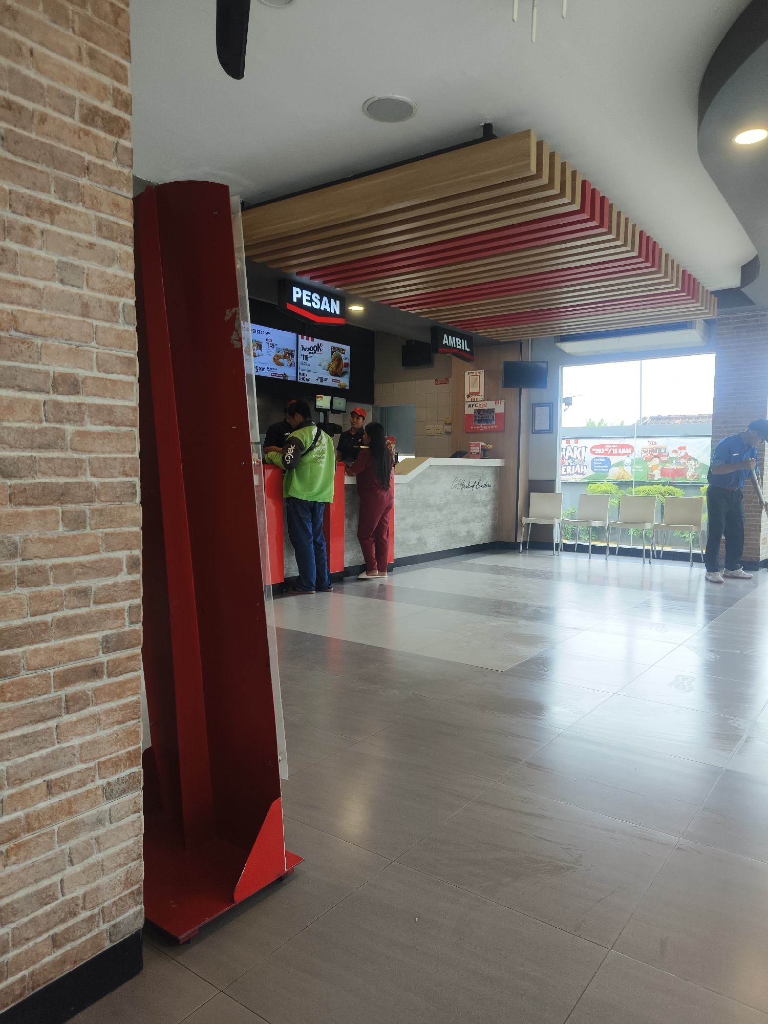 The image depicts the interior of a KFC restaurant in Denpasar, Indonesia. In the foreground, a red promotional stand is positioned against a brick wall. Beyond this, a counter area is visible with several individuals. A person wearing a green jacket with "gojek" branding is interacting with staff at the counter. To the right of the counter, a person in a red uniform is also present. Further to the right, a man in a blue uniform is sweeping the floor. The environment includes a large window offering a view of the exterior, seating chairs, and a modern ceiling design with wooden and red slats. The floor is tiled, and the overall lighting suggests daytime.