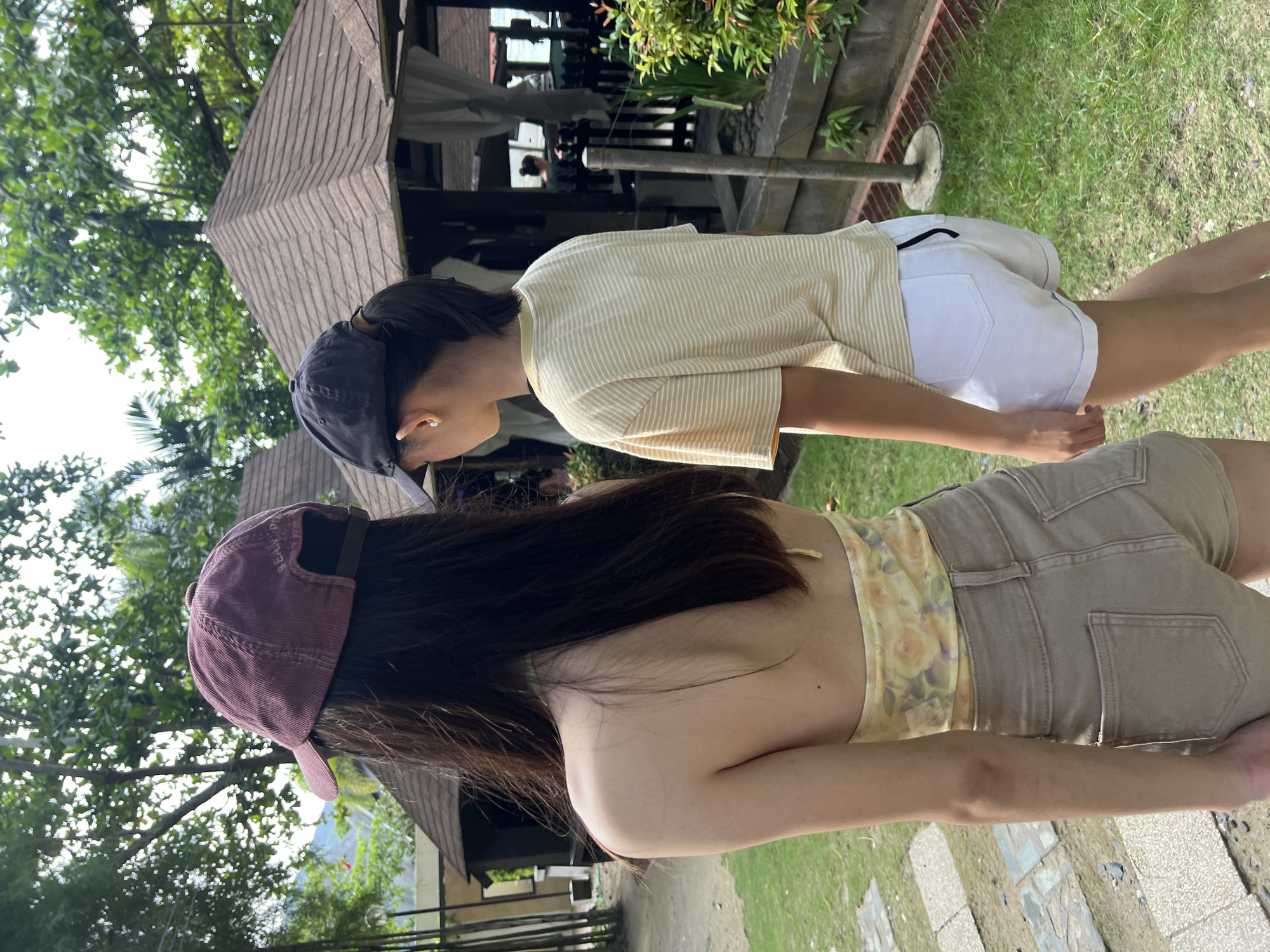 Two individuals are standing outside in a grassy area. The person in the foreground has long, dark hair and is wearing a mauve-colored baseball cap, a yellow floral top, and beige shorts. The other person has short, dark hair and is wearing a dark blue baseball cap, a yellow striped t-shirt, and white shorts. Behind them are some buildings, trees, and grass. The location is in Banaybanay, Philippines.
