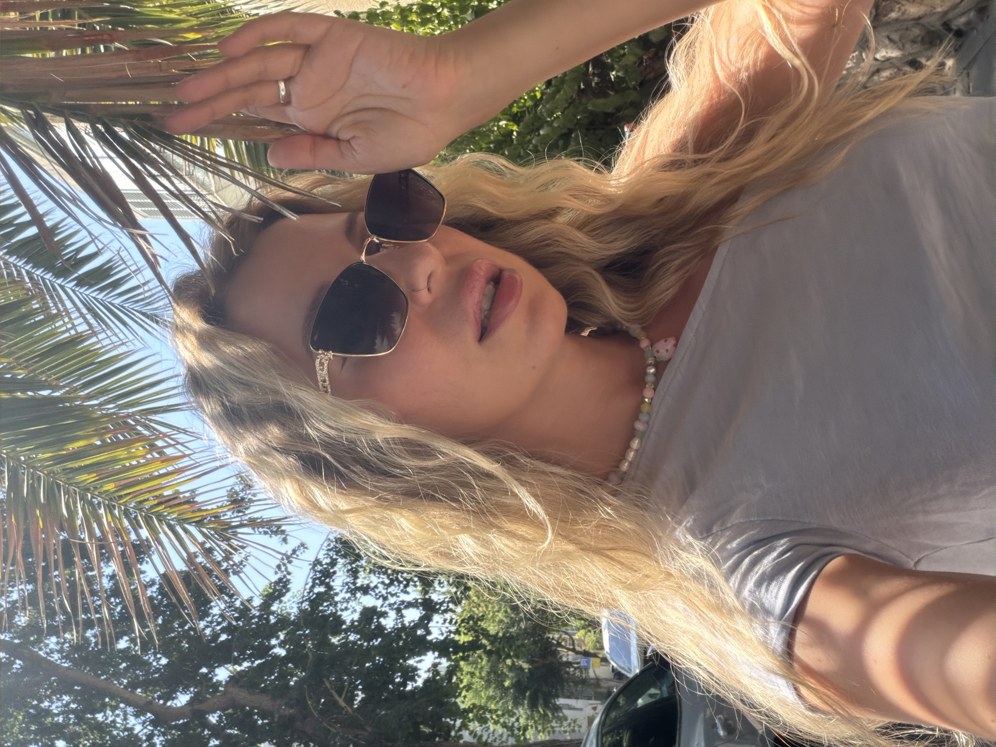 An outdoor, eye-level, medium shot captures a blonde woman in sunglasses and a light gray t-shirt, posing outdoors. She is partially obscured by palm fronds casting shadows on her. The woman is wearing a pearl necklace with a pink pendant. Her right hand is raised, touching her hair and sunglasses, with a ring visible on her ring finger. In the background, trees, buildings, and a parked car are visible, suggesting an urban environment, likely in Bat Yam, Israel, given the presence of palm trees and Mediterranean architecture.