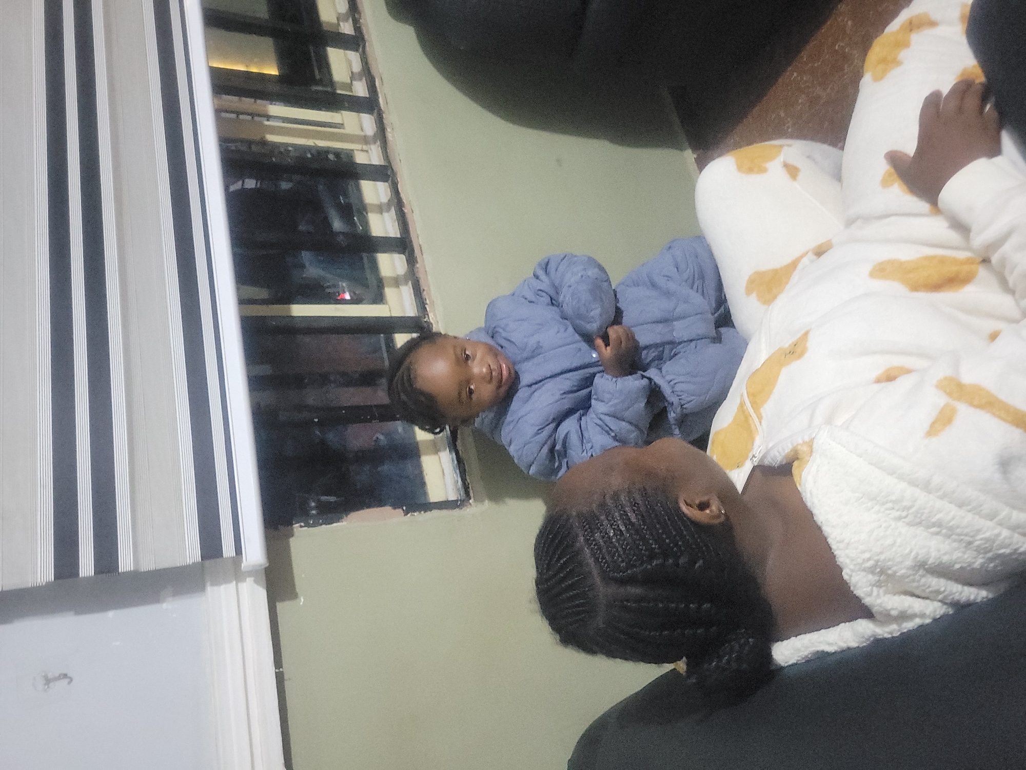 Two individuals are situated within an indoor environment, likely a residential setting. A young child, dressed in a blue, quilted jacket, is positioned near a window. An adult lies nearby, wearing a white garment adorned with tan patterns. The adult has braided hair and a fair complexion. The setting features a window with Venetian blinds and a view of a dark cityscape. The city and exact location are unspecified.