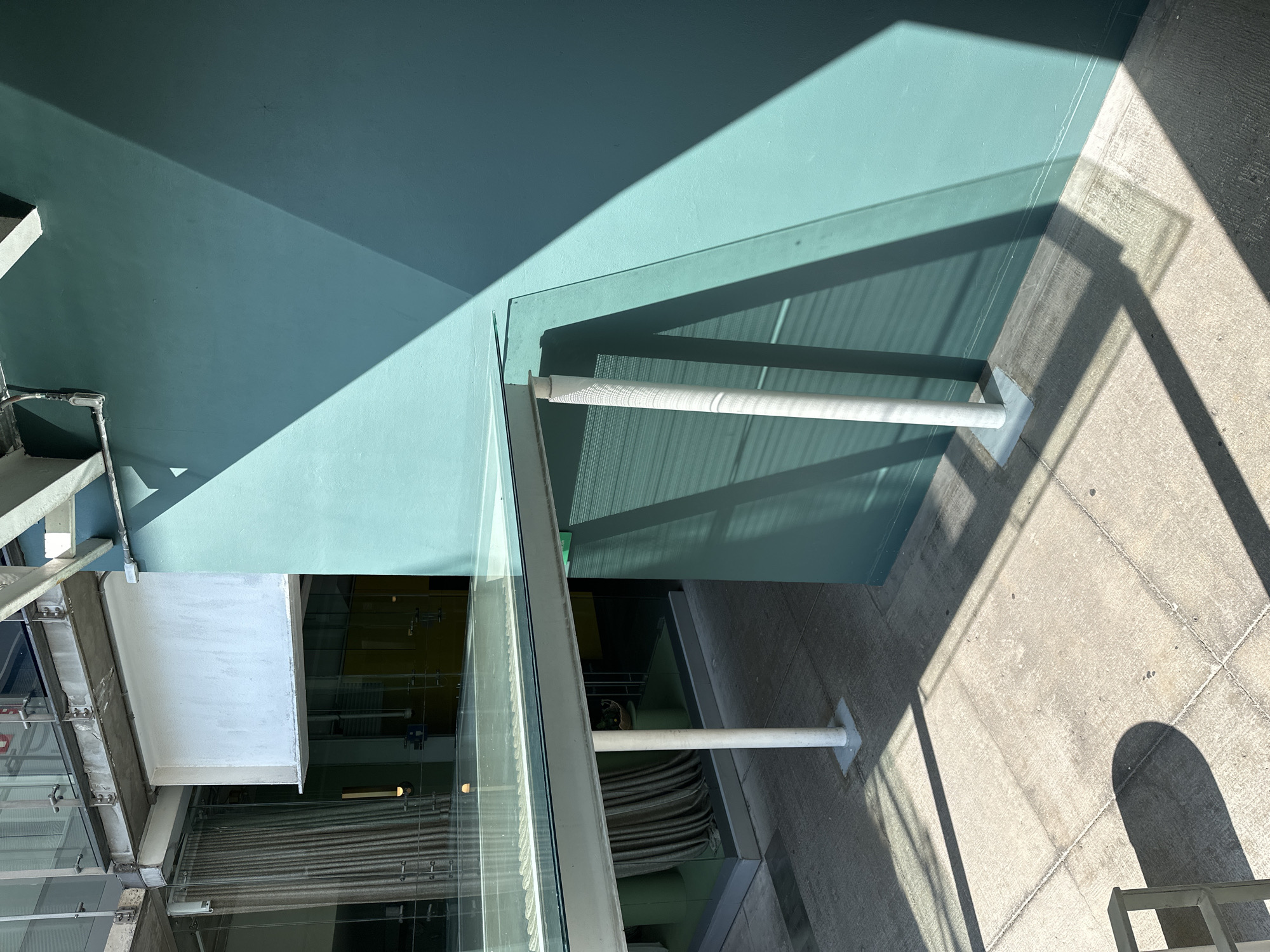 The image depicts a modern architectural scene, possibly in Mexico City, characterized by a blend of concrete, glass, and steel elements. The dominant visual feature is a teal-painted overhead structure, which casts a stark shadow on the adjacent concrete ground. The structure appears to be an awning or covering, possibly part of a building facade. Supporting the awning are white cylindrical posts. A glass wall runs parallel to the awning, reflecting the surrounding environment and revealing glimpses of an interior space with yellow and gray elements. A portion of a steel staircase is visible in the left corner. The overall composition suggests a contemporary urban setting, emphasizing the interplay of light and shadow on geometric forms. The concrete surface shows wear and the seams of the sections that make it up.