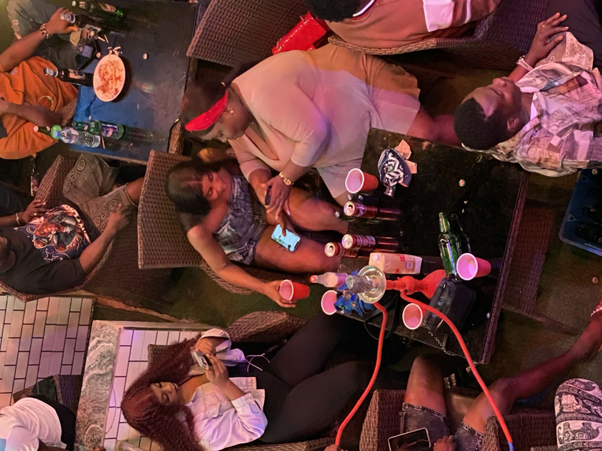 This media file captures a group of people socializing at what appears to be an outdoor lounge or restaurant. The scene is set at night, with warm ambient lighting creating a lively and relaxed atmosphere. The subjects are diverse, including men and women dressed in casual attire.
Several objects are visible on the tables, suggesting a gathering focused on drinks and socializing. These include bottles of various beverages, red plastic cups, and a shisha pipe, indicating that smoking shisha is part of the activity. There's also a plate with food, possibly a shared snack. One individual is using a smartphone, while another is holding one up.
The people are seated on wicker chairs around low tables, and the ground beneath them is a mix of grass and tiled areas. The overall mood seems to be one of enjoyment and camaraderie, with people engaged in conversation and relaxation. The presence of a shisha pipe and the attire suggest a contemporary social setting. While specific textual information is limited, the visual cues point towards a casual evening hangout in Obosi, Nigeria.