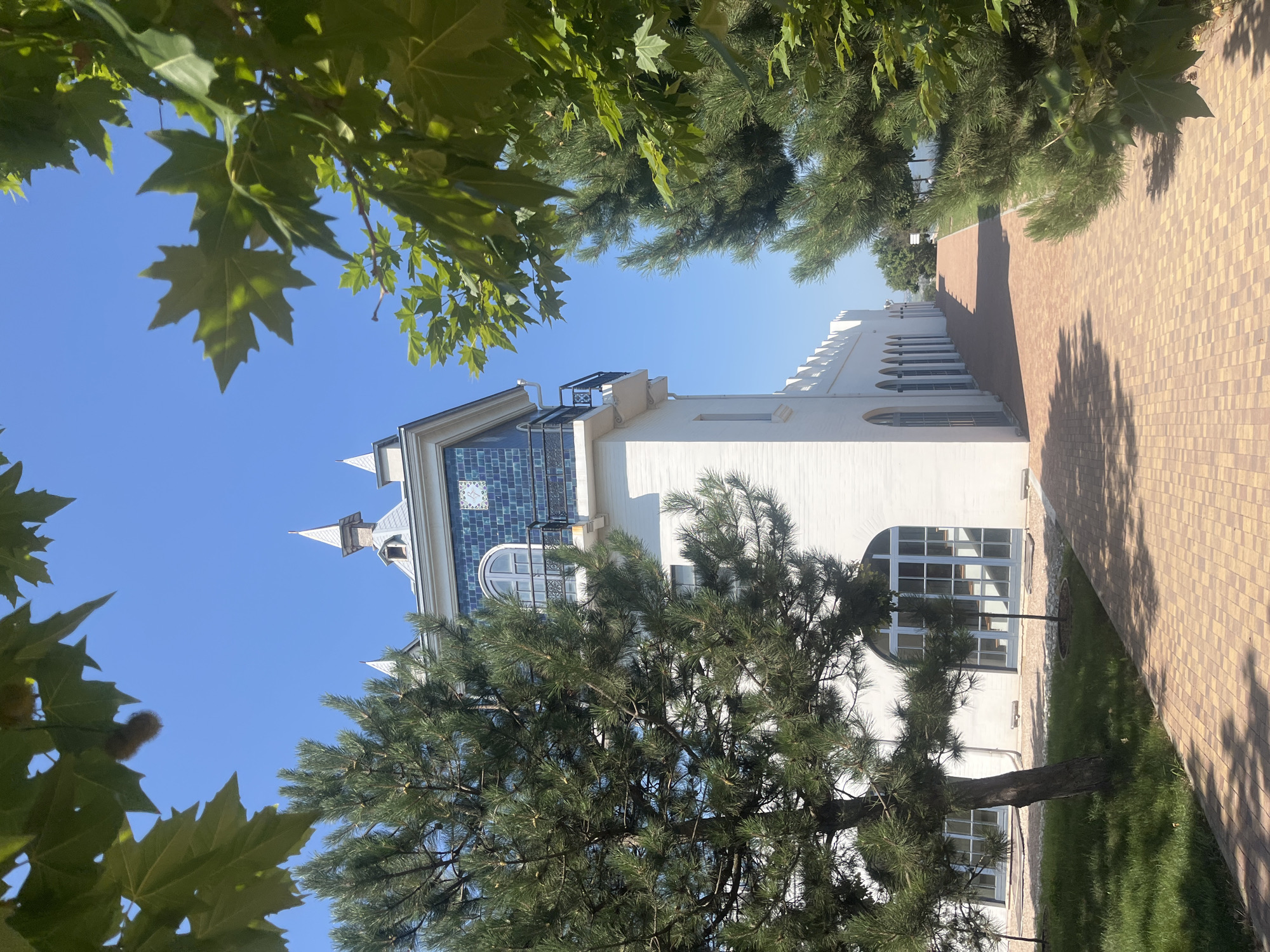 The image depicts a paved walkway leading past a white building with a tiled tower. The building features arched windows and a crenellated roofline. Large pine trees and leafy trees frame the scene, casting shadows on the walkway. The sky is clear and blue. The location is identified as Peresyp, Russia. There are no people or visible actions in the image.