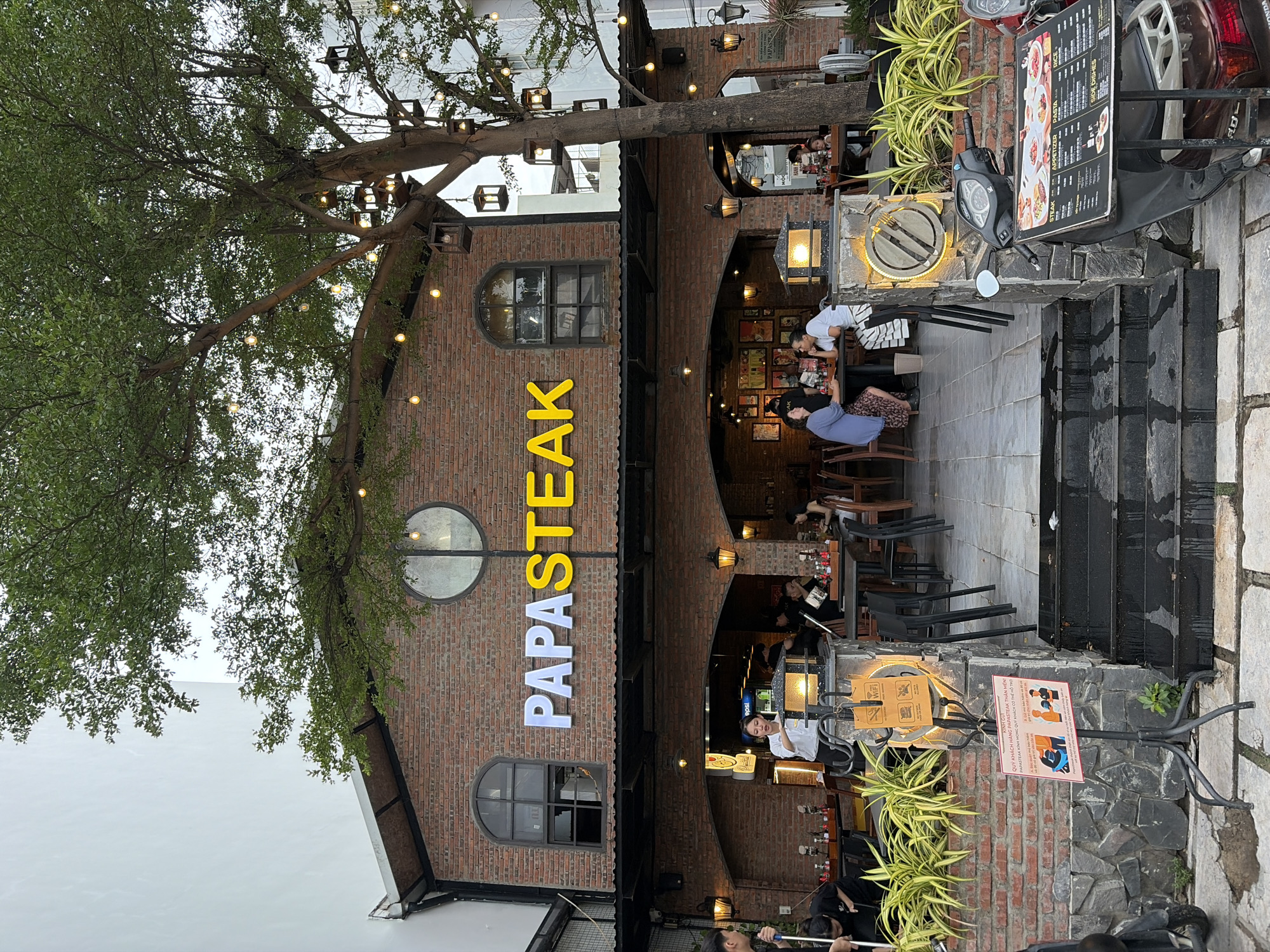 The image depicts the exterior of a restaurant named "PAPASTEAK" in Đà Nẵng, Vietnam, at dusk or overcast conditions. The building is constructed with red brick and features arched entrances and windows. A large tree with decorative lights and lanterns is positioned to the right of the entrance, providing shade and ambiance.
Several patrons are seated at tables both inside and outside the restaurant. Some staff members are visible, including one person sweeping the entrance area. A menu board is placed on a scooter parked to the right of the steps leading into the restaurant. A sign with Vietnamese text and pictograms indicating rules for customers is mounted on a stand near the steps. The overall scene suggests a casual dining establishment with outdoor seating, set against a backdrop of brick architecture and lush greenery.