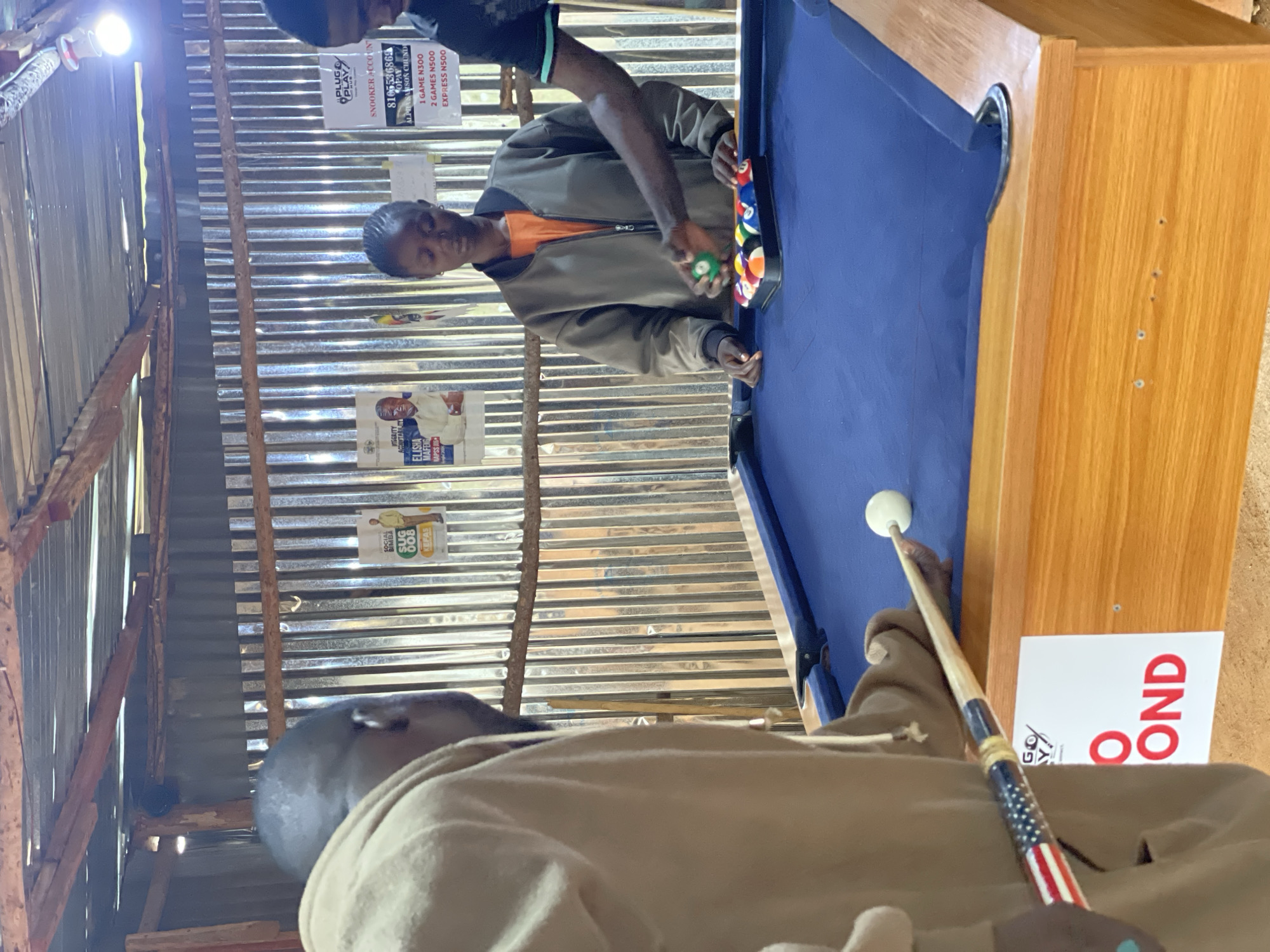 In Durumi, Nigeria, a group of individuals are gathered around a blue pool table in an open-air establishment with a corrugated metal roof. The walls are also constructed of corrugated metal. A man in a tan hoodie is holding a pool cue decorated with the American flag. In the center of the frame, a young woman in a grey jacket over an orange shirt is placing a green pool ball into a rack of multi-colored pool balls. To her right, another man's arm is visible, also interacting with the pool balls. Behind the people, posters are affixed to the metal wall. One poster appears to be an election advertisement, while another displays pricing information for "PLUG & PLAY" snooker games. The floor of the establishment is unpaved earth.