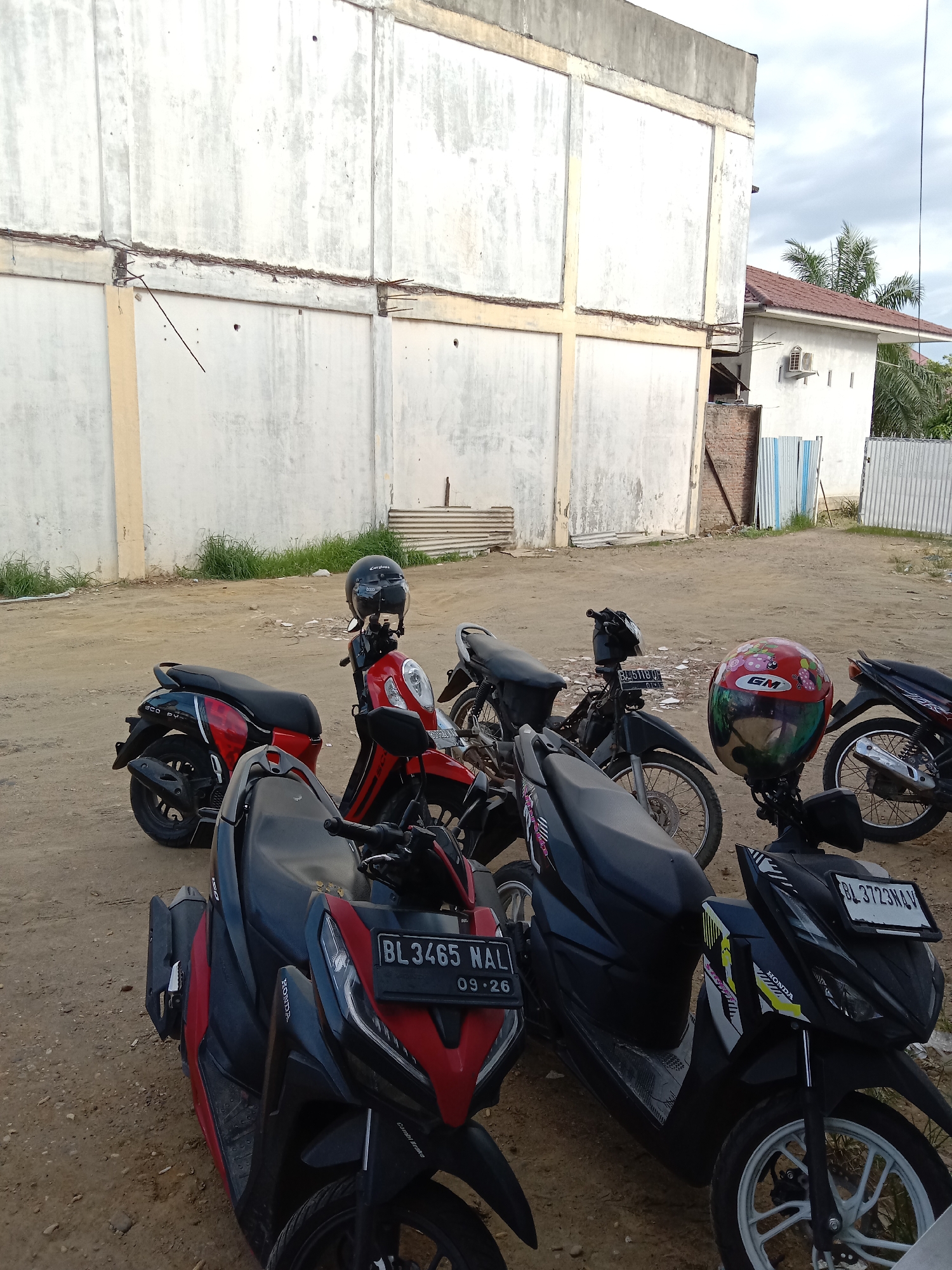 The image shows a parking area in Lhokseumawe, Indonesia, filled with motorbikes. A large, unfinished white concrete building is visible in the background. In front of it, there are seven motorbikes parked on a sandy surface. Helmets rest on some of the bikes. License plates are visible on the front motorbikes, reading "BL 3465 NAL" and "BL 3723 NAV". To the right of the concrete structure, a smaller white building with an air conditioning unit and a red roof is partially visible behind a fence. The sky is overcast. The overall scene suggests a typical parking area adjacent to a construction site or commercial building in an urban area of Lhokseumawe.